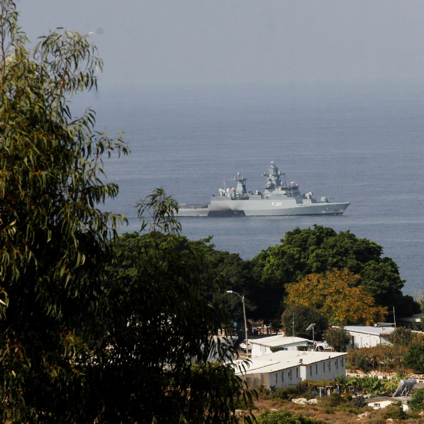 در تصویر، یک کشتی جنگی در نزدیکی ساحل دیده می‌شود که در دریا شناور است. این کشتی دارای طراحی مدرن و تجهیزات نظامی است. در پیش‌زمینه، تعدادی درخت و ساختمان‌های کوچک دیده می‌شود، و پس‌زمینه نمایی از دریا و آسمان روشن دارد. تضاد بین رنگ‌های سبز درختان و آبی دریا جلوه‌ای زیبا به تصویر بخشیده است.