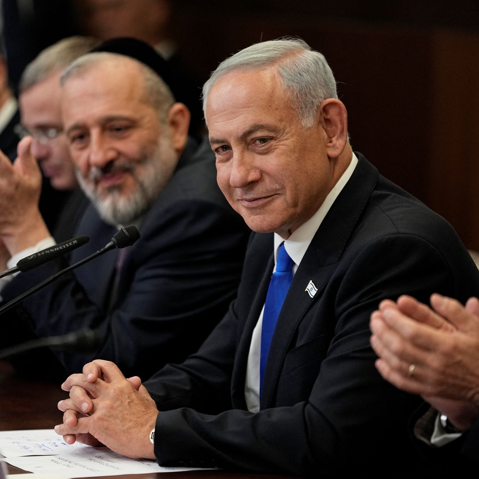 L'image montre un groupe de personnes assises autour d'une table, probablement lors d'une réunion. Au centre se trouve un homme en costume avec une cravate bleue, qui semble écouter attentivement et sourire. Les autres personnes autour de lui applaudissent et affichent des expressions positives. L'environnement est formel, avec une décoration sobre.