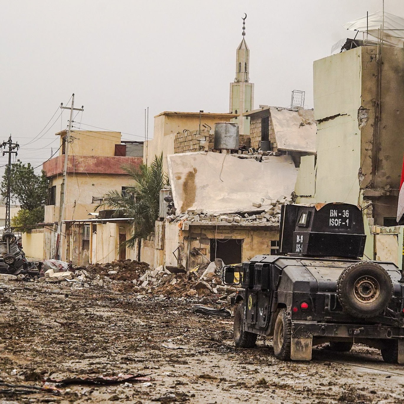 الصورة تظهر مشهداً من منطقة متضررة بسبب نزاع أو حرب. هناك الكثير من الخراب في المباني المحيطة، حيث تبدو العديد منها مدمرة أو شبه مدمرة. في منتصف الصورة، توجد مركبة عسكرية، يبدو أنها تُستخدم في عمليات الأمن أو الاستجابة الطارئة. تلوح في الأفق مئذنة مسجد، مما يشير إلى وجود مجتمع محلي. الشوارع تبدو مليئة بالتراب والحطام، مما يعكس آثار الدمار الذي لحق بالمنطقة.