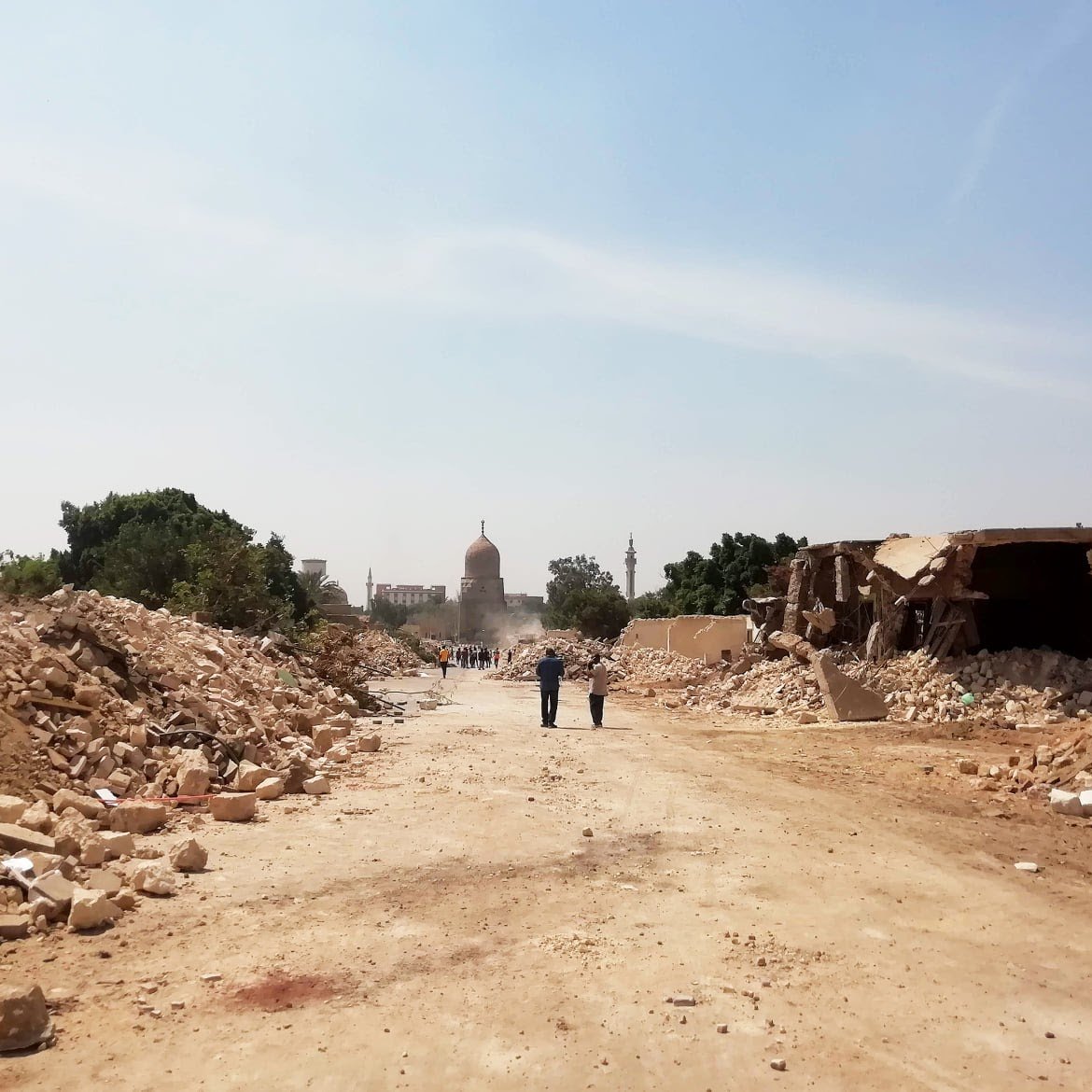 L'immagine mostra una strada sterrata, circondata da macerie e detriti. Sullo sfondo si intravede un edificio con una cupola, probabilmente una moschea o un monumento, avvolto da polvere. La scena trasmette un senso di devastazione, con alberi e strutture danneggiate ai lati della strada. Alcune persone sono visibili in lontananza, mentre il cielo appare chiaro e luminoso.