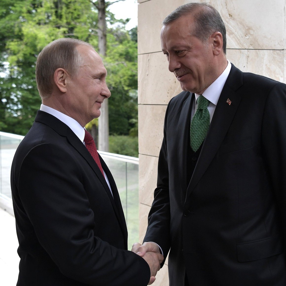 L'image montre deux hommes en costume, se serrant la main. L'un d'eux porte un costume noir avec une cravate rouge, tandis que l'autre est également en costume noir, mais avec une cravate verte. Ils se tiennent à l'extérieur, devant un fond de verdure et d'architecture moderne. L'atmosphère semble conviviale et formelle.