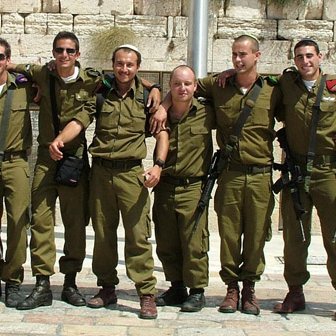 The image features a group of eleven soldiers standing closely together, wearing olive green military uniforms and boots. They appear to be in a celebratory mood, with smiles and casual poses. The background suggests a stone wall, possibly the Western Wall in Jerusalem, indicating a significant location. The soldiers carry some equipment, hinting at their military roles. Overall, the image conveys camaraderie and a sense of accomplishment among the group.
