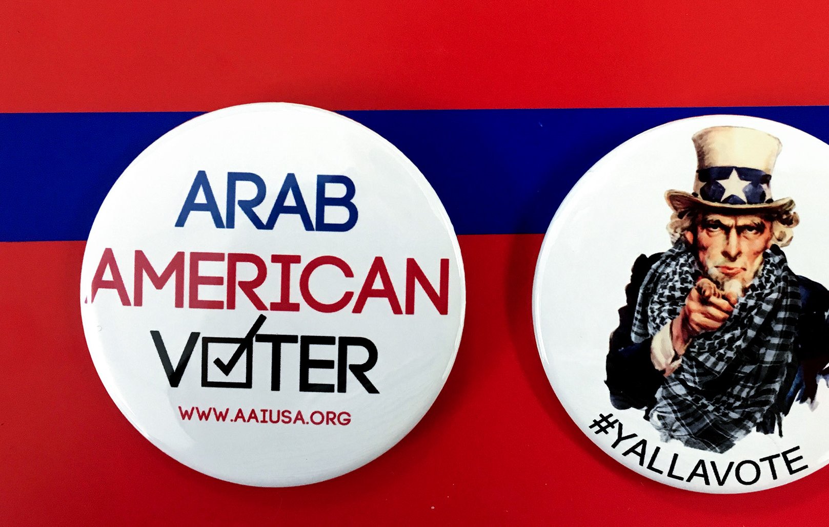 الصورة تحتوي على زرين دائرين. الزر الأول مكتوب عليه "ARAB AMERICAN VOTER" مع علامة التحقق، مما يدل على التصويت للمواطنين العرب الأمريكيين. الزر الثاني يظهر صورة لشخص يرتدي قبعة تحمل نجمة، وهو رمز معروف في الثقافة الأمريكية، مع عبارة "#YALLAVOTE" التي تدعو الناس للتصويت. الخلفية حمراء مع شريط أزرق، مما يضيف طابعاً احتفالياً للموضوع.
