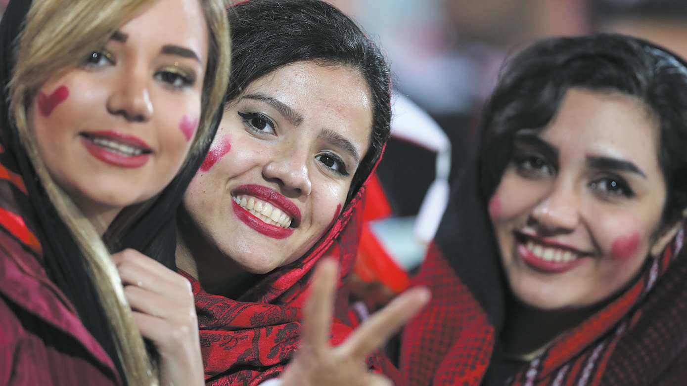 L'image montre trois femmes souriantes, vêtues de vêtements dans des tons sombres, avec des écharpes ou des foulards sur la tête. Chacune d'elles a des cœurs rouges dessinés sur les joues, ce qui suggère qu'elles célébrent un événement sportif ou une occasion festive. Elles affichent des expressions de joie et de camaraderie, et l'une des femmes montre un signe de paix avec ses doigts. L'arrière-plan est flou, mais on peut discerner une ambiance festive.
