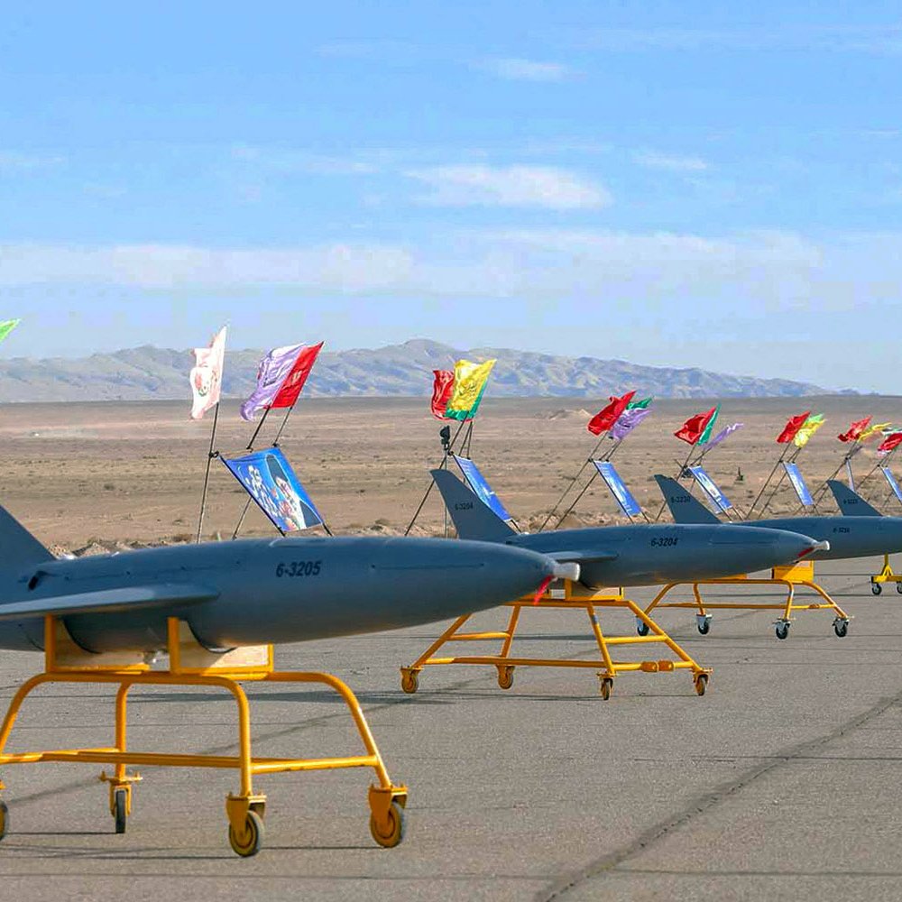 La imagen muestra una fila de drones en una pista desértica. Estos drones están montados sobre soportes amarillos y llevan banderas de diferentes colores. Al fondo, se puede ver un paisaje montañoso y un cielo despejado. La escena parece estar relacionada con una exhibición o un evento militar.