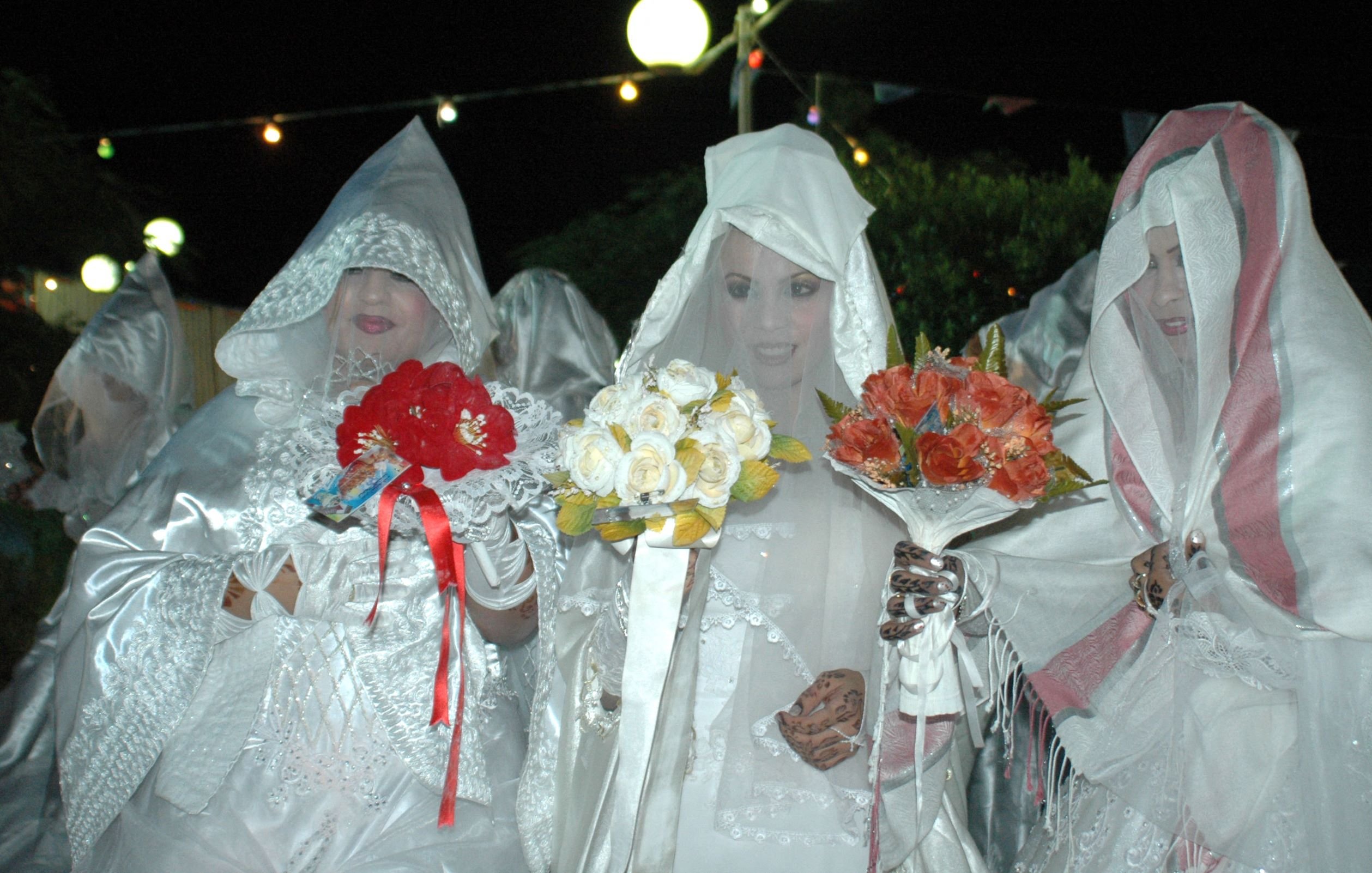 L'immagine mostra un gruppo di donne vestite con abiti bianchi e coperti da veli, che formano un'atmosfera suggestiva. Alcune di loro tengono in mano mazzi di fiori colorati, come rose rosse e fiori bianchi. L'illuminazione è soffusa, creando un'atmosfera notturna, e sullo sfondo si possono vedere luci decorative. Le donne sembrano partecipare a un evento particolare, probabilmente una celebrazione o un rituale tradizionale.