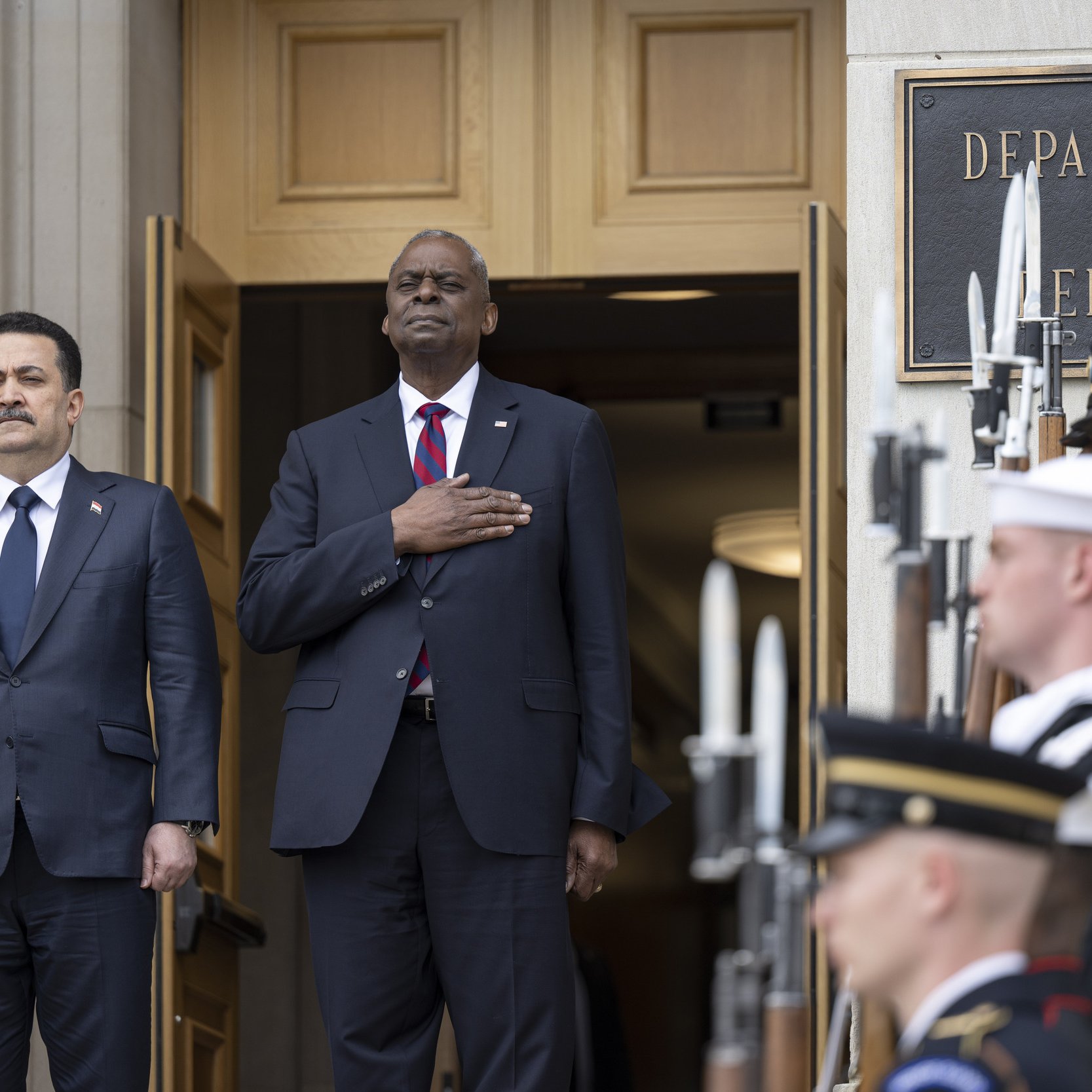 L'immagine mostra due uomini in piedi davanti all'entrata di un edificio governativo, probabilmente il Dipartimento della Difesa. Uno di loro indossa un abbigliamento formale con un completo scuro e una cravatta, mentre l'altro ha il braccio destro sul cuore, indicando un gesto di rispetto o di onore. Dietro di loro, ci sono diversi membri delle forze armate in uniforme, schierati con armi in mano, pronti a rendere onore. L'atmosfera è solenne e pomposa, suggerendo un evento ufficiale o una cerimonia.