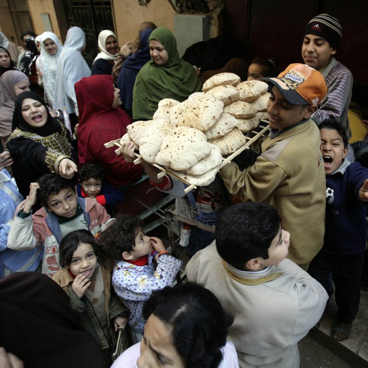 L'image montre une scène animée dans une rue, probablement dans un pays arabe. Un jeune garçon porte un plateau chargé de pains, entouré d'une foule de personnes, comprenant des adultes et des enfants. On peut voir des visages souriants et des expressions variées, ce qui donne une impression de convivialité et d'effervescence. Les gens semblent attendre pour acheter du pain, et la diversité des vêtements suggère une ambiance culturelle riche. L'image capte un moment de vie quotidienne, centré sur l'échange et la nourriture.