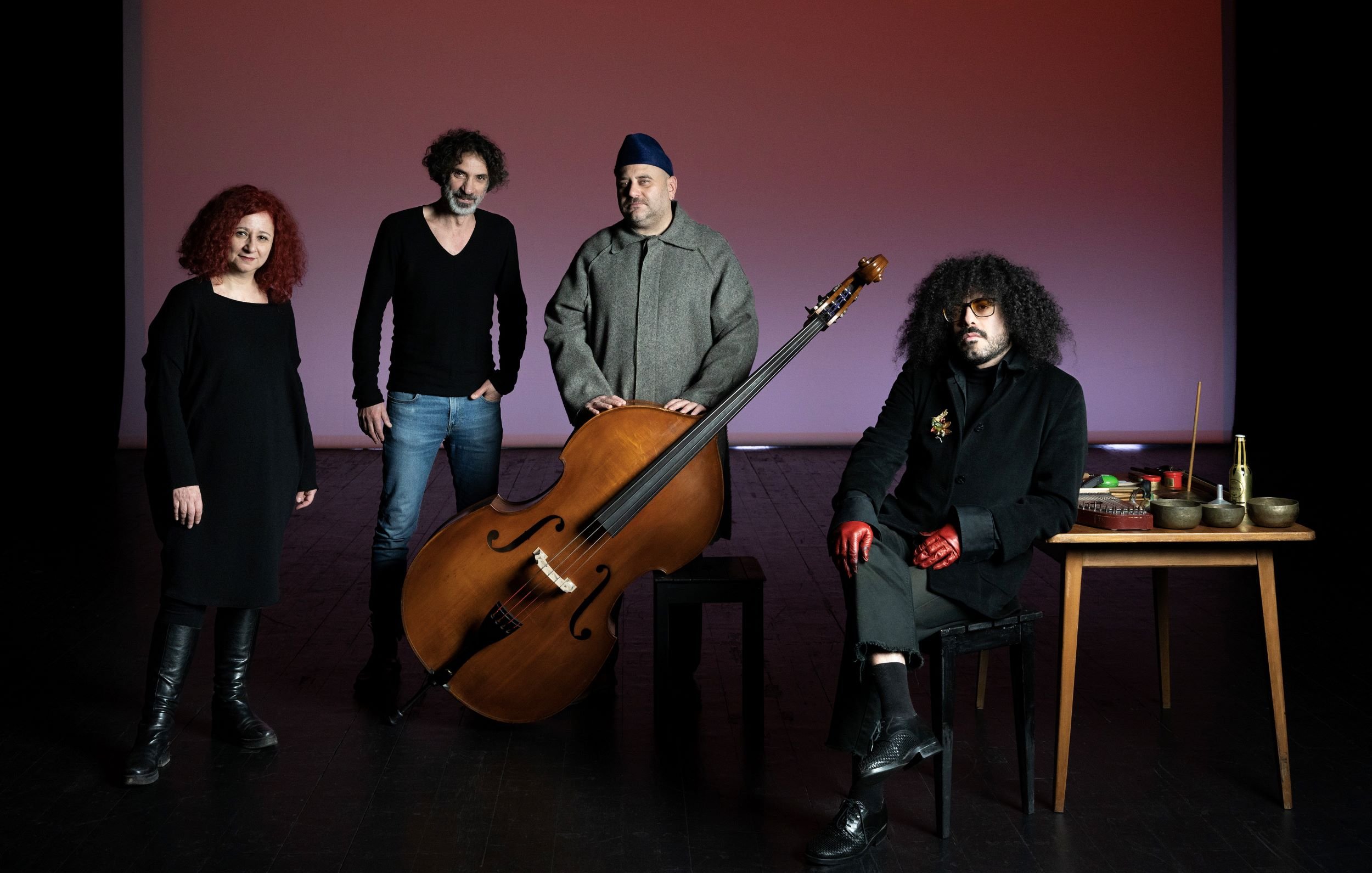 L'image présente un groupe de quatre personnes posant ensemble sur scène. Au centre, il y a un grand instrument de musique, probablement un instrument à cordes, qui attire l'attention. À gauche, une femme avec des cheveux roux bouclés se tient avec une expression sérieuse. À côté, un homme avec des cheveux frisés et un pull noir semble également concentré. Au centre, près de l'instrument, un homme porte une tenue grise et un couvre-chef. Enfin, à droite, un autre homme avec des lunettes et des gants rouges est assis à une table avec divers objets, ayant l'air intrigant. L'éclairage de la scène crée une ambiance artistique et captivante.