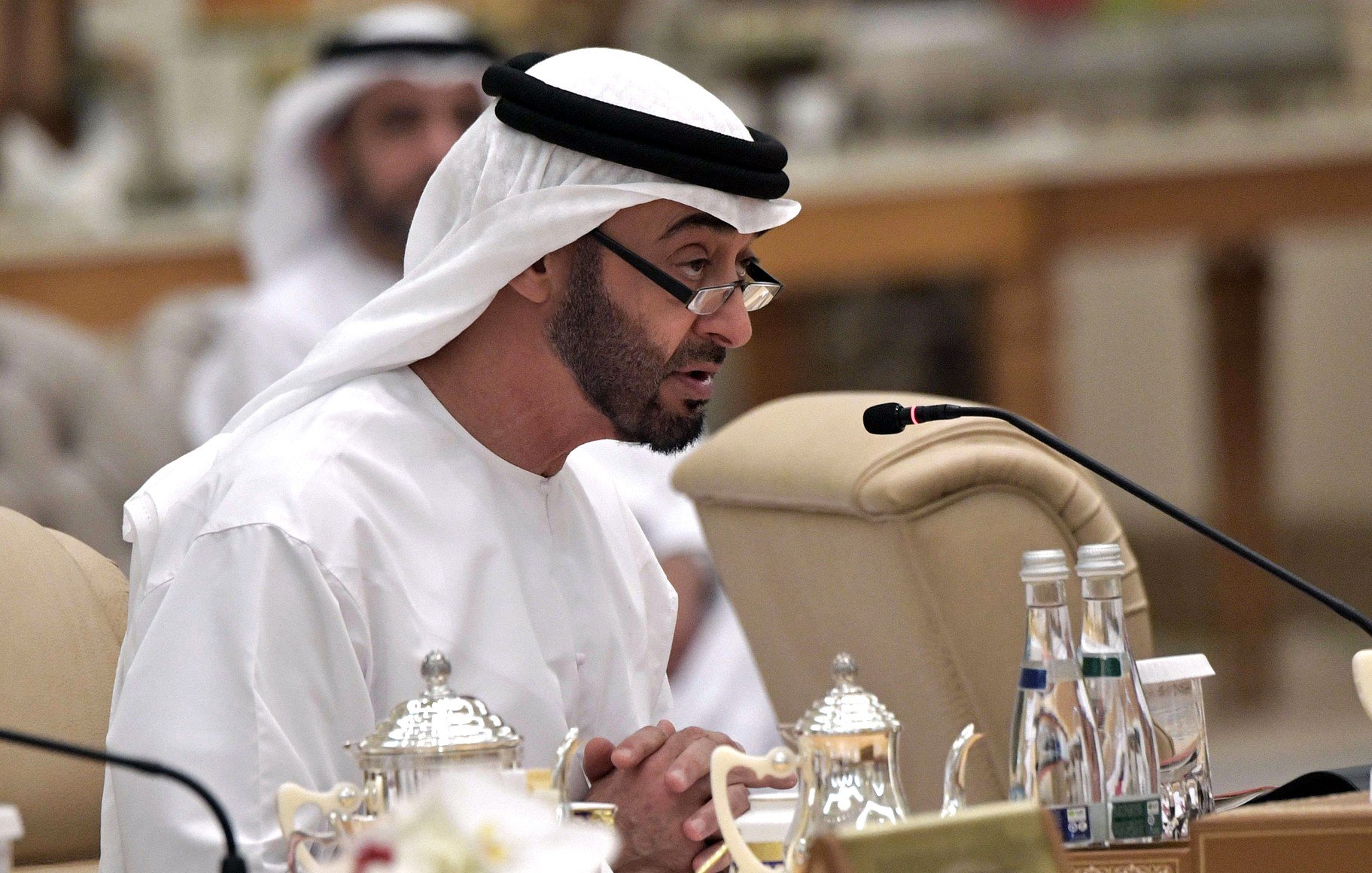 تظهر في الصورة شخصية ترتدي الزي التقليدي الإماراتي، بما في ذلك الغترة والعقال. يبدو أنه في وضع يتحدث خلال اجتماع أو جلسة حوارية، حيث يوجد ميكروفون أمامه. في الخلفية، يمكن رؤية بعض الأشخاص الآخرين المشاركين في الاجتماع. الطاولة مُزينة بأدوات الشاي وبعض الزهور، مما يضيف لمسة من الأناقة والضيافة.