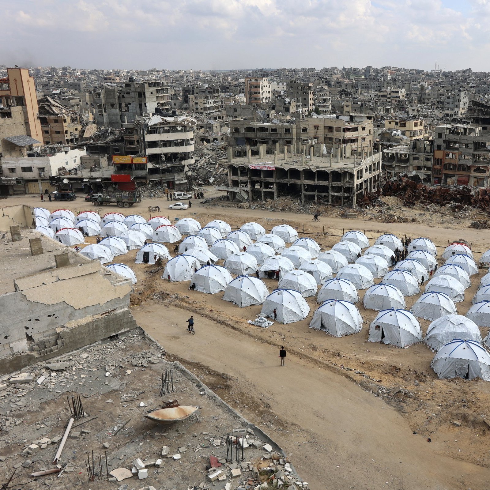 این تصویر نمایی از یک منطقه تخریب‌شده را نشان می‌دهد که در آن چادرهای سفید رنگ به‌عنوان محل اسکان موقت قرار گرفته‌اند. در پشت این چادرها، ساختمان‌های ویران دیده می‌شوند که نشان‌دهنده آثار جنگ یا بلایای طبیعی هستند. زمین خاکی و خالی با چندین چادر در صف‌های منظم، حس نیاز و بحران را منتقل می‌کند. آسمان نیز ابری به نظر می‌رسد که به فضای تصویر افزوده است.