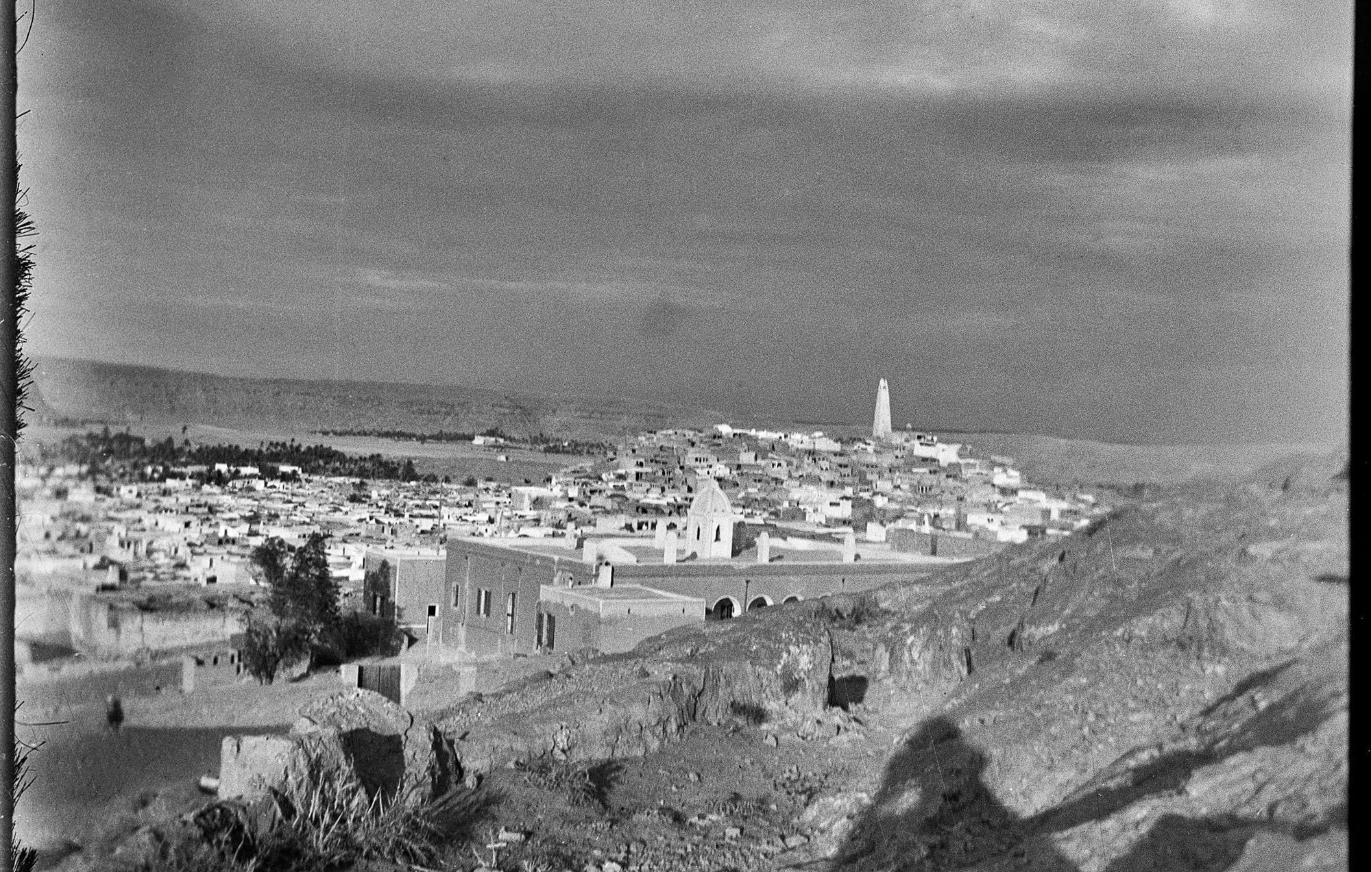 L'image montre un paysage urbain, probablement d'une ville historique. On peut voir des maisons blanches et des structures architecturales qui s'étendent à travers la vallée. À l'arrière-plan, un minaret s'élève, suggérant une présence religieuse. Le ciel semble nuageux, ce qui ajoute une ambiance dramatique à la scène. Le terrain est rocailleux au premier plan, et l'éclairage donne une impression de nostalgie, typique des photographies anciennes.
