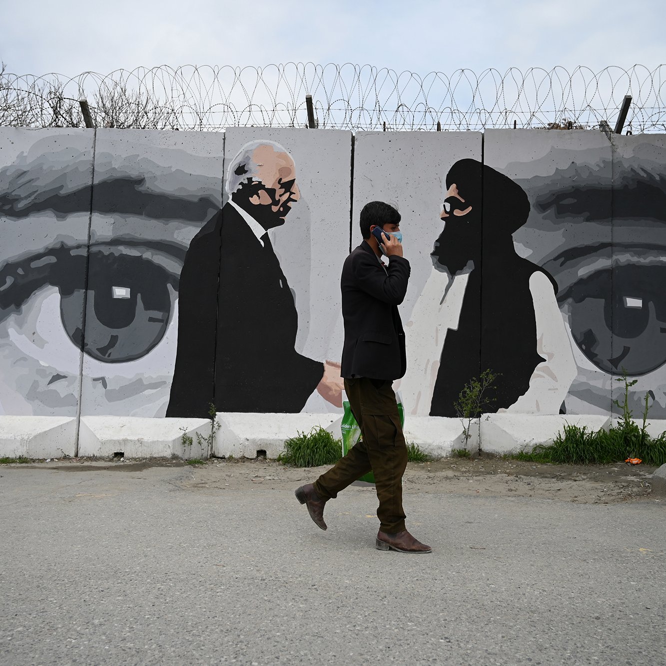 La imagen muestra a un hombre caminando por una calle, mientras habla por teléfono. Detrás de él hay un mural que representa dos figuras en blanco y negro, aparentemente en un diálogo. El mural incluye grandes ojos que observan la escena. Además, se pueden ver alambres de púa en la parte superior del muro que sostiene el mural, lo que sugiere un ambiente de tensión o seguridad. La iluminación es natural, indicando que es de día.