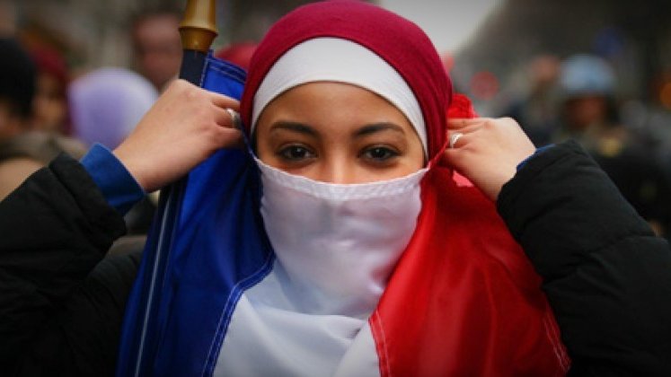 L'image montre une personne portant un hijab qui utilise un drapeau français comme écharpe. Elle a un regard déterminé et semble faire partie d'une manifestation ou d'un rassemblement. L'arrière-plan montre une foule, ce qui suggère un contexte collectif et engagé. Les couleurs du drapeau français, bleu, blanc et rouge, sont bien présentes, symbolisant un élan de patriotisme.