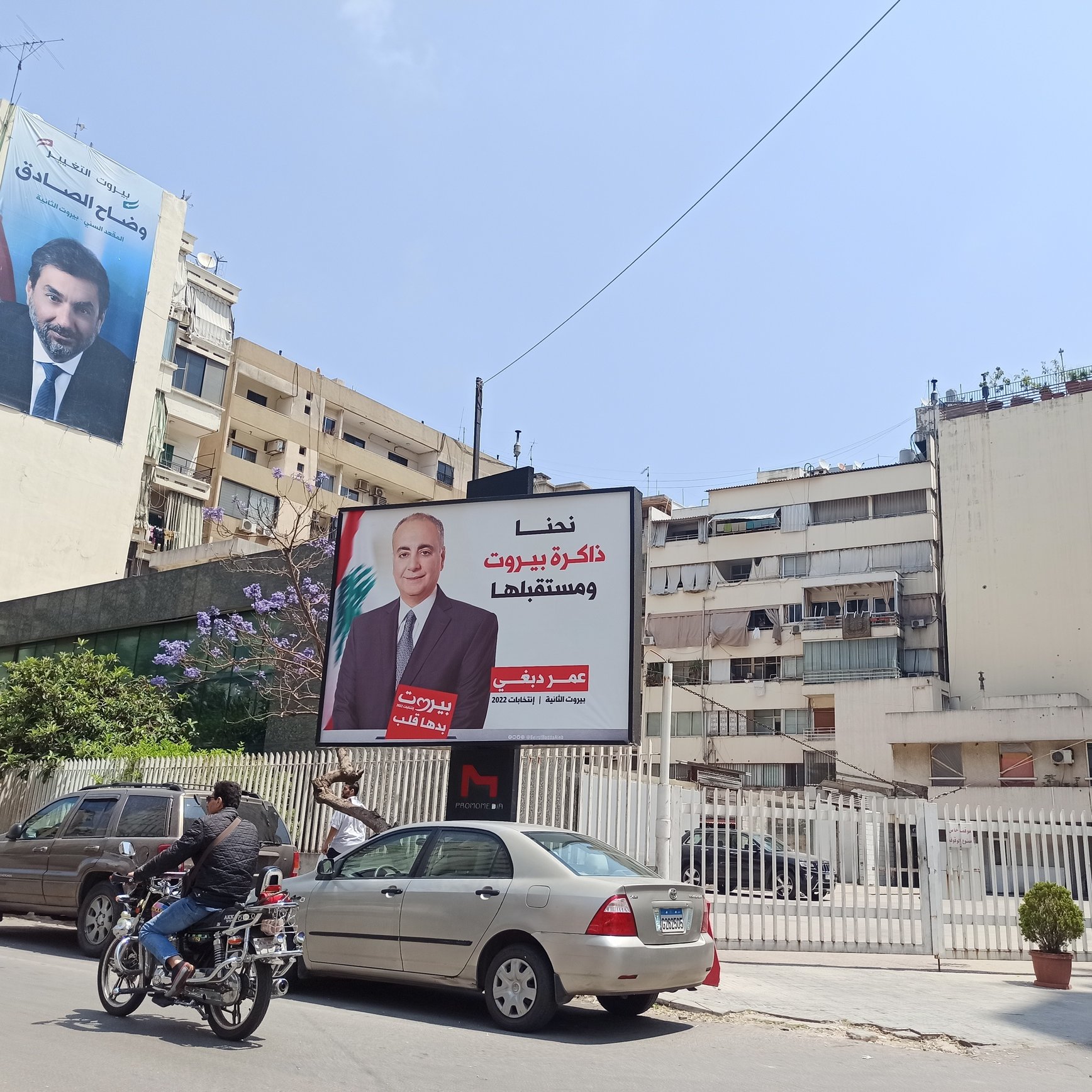 L'image montre une rue animée avec des voitures et des motos en circulation. À gauche, il y a une grande affiche sur un bâtiment, probablement une campagne politique, mettant en avant une personne, avec des textes en arabe. À côté, un autre bâtiment est visible, avec une façade claire et quelques plantes qui ajoutent une touche de verdure à l'environnement urbain. Le ciel est bleu, ce qui donne une ambiance ensoleillée à la scène.
