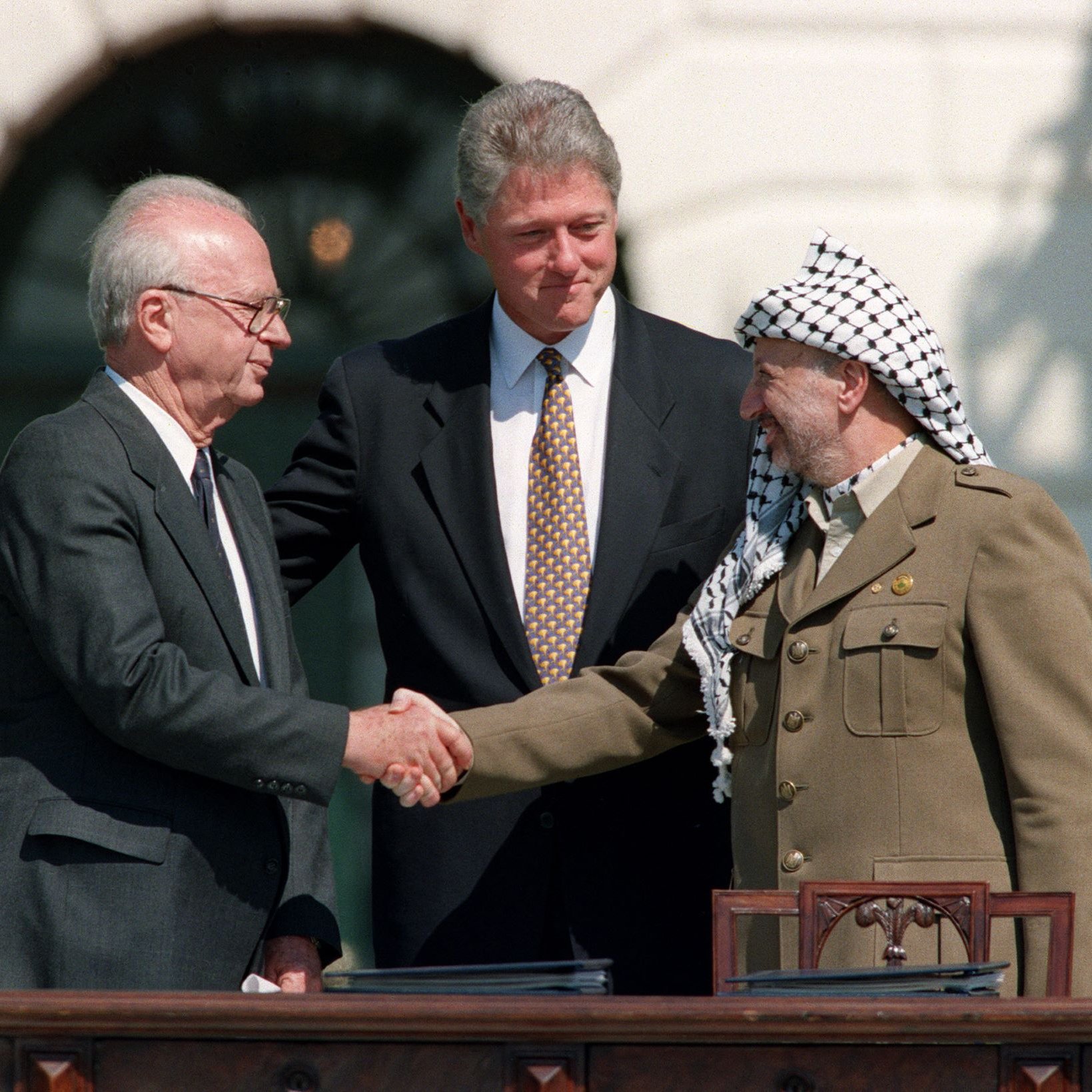L'image montre trois hommes se serrant la main lors d'un événement important. À gauche, un homme âgé avec des lunettes, à droite, un homme en costume militaire, et au centre, un homme en costume noir qui semble jouer un rôle de médiateur. Ils se tiennent sur un balcon ou une terrasse, avec un décor architectural en arrière-plan. Cette scène évoque un moment symbolique de paix ou de négociation entre des parties en conflit.