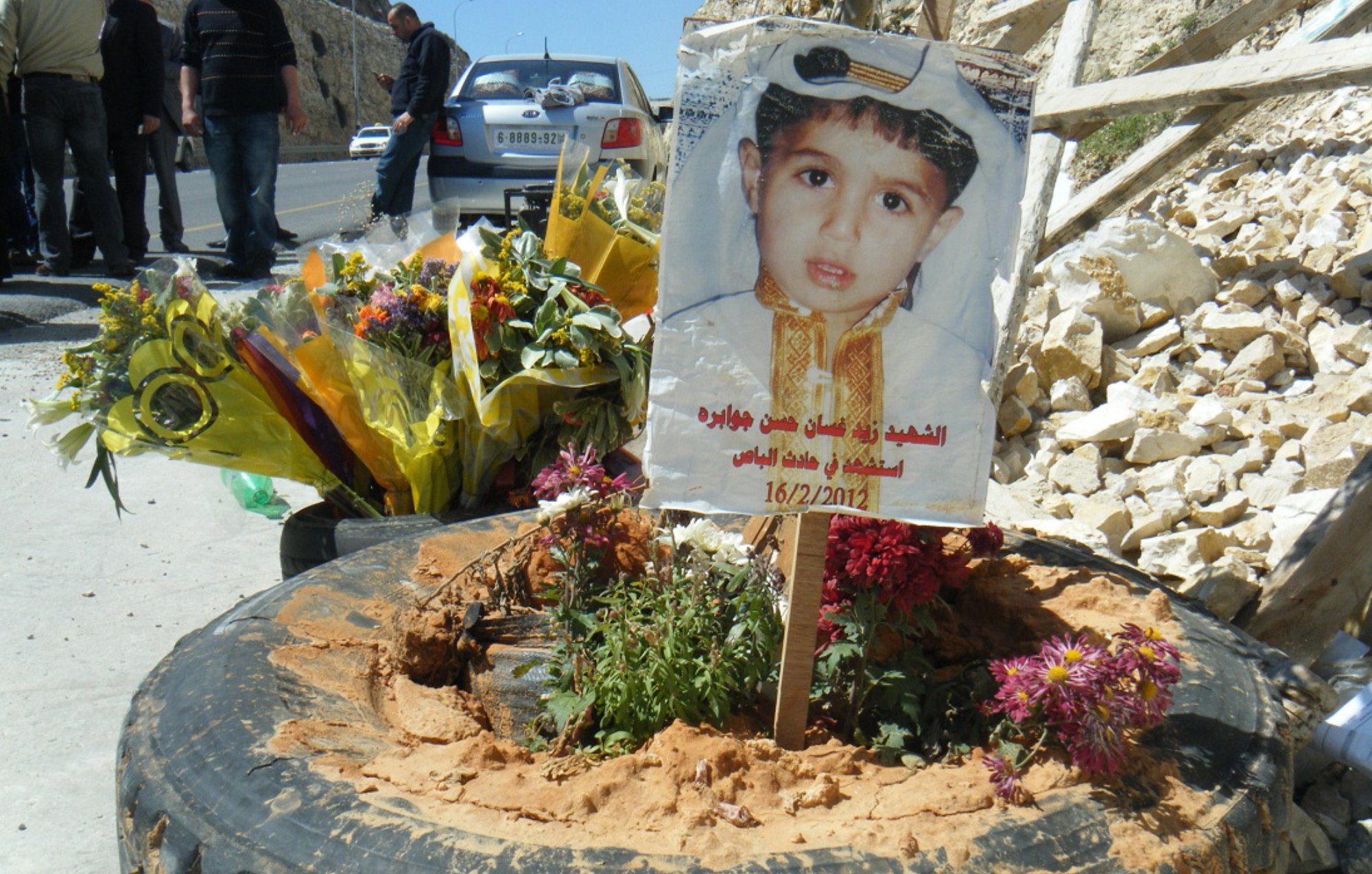 L'immagine mostra un tributo commemorativo collocato su un terreno. Al centro, c'è un cartello che presenta un ritratto di un bambino, adornato con un abbigliamento tradizionale. Attorno al cartello ci sono fiori freschi e piante, posizionati in una gomma di automobile. Il tutto è circondato da un ambiente duro, con rocce visibili e alcune persone in fondo che sembrano partecipare a una commemorazione. La data sul cartello indica un giorno significativo, possivelmente legato alla memoria del bambino.