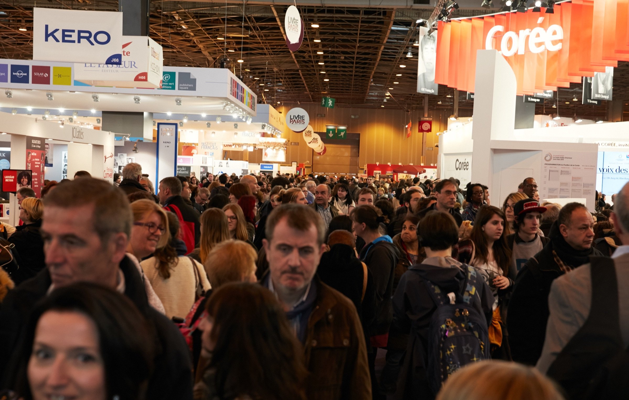 L'image montre une foule dense de personnes rassemblées dans un espace d'exposition, probablement lors d'un événement ou d'une foire. On peut voir des stands avec des bannières et des affiches en arrière-plan, créant une atmosphère dynamique. Les gens sont variés en termes de styles vestimentaires et d'âges, indiquant un mélange diversifié de visiteurs. L'éclairage est lumineux, mettant l'accent sur l'activité animée qui se déroule dans ce lieu.