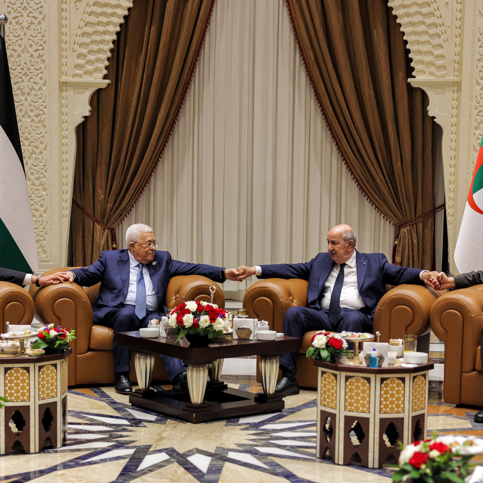 تظهر الصورة مجموعة من الأشخاص seated في غرفة مزينة بشكل أنيق، حيث يجلس أربعة رجال على أرائك مريحة. بينهم أجواء من التعاون والود، إذ يتبادلون الحديث مع قيامهم بتشابك الأيادي. في الخلفية، توجد أعلام فلسطين والجزائر، ما يشير إلى وجود موضوع يتعلق بالتعاون العربي أو القضايا السياسية بين البلدين. الطاولات أمامهم مزينة بتنسيقات من الزهور ومشروبات. الأجواء تبدو رسمية ولكن ودية في الوقت نفسه.