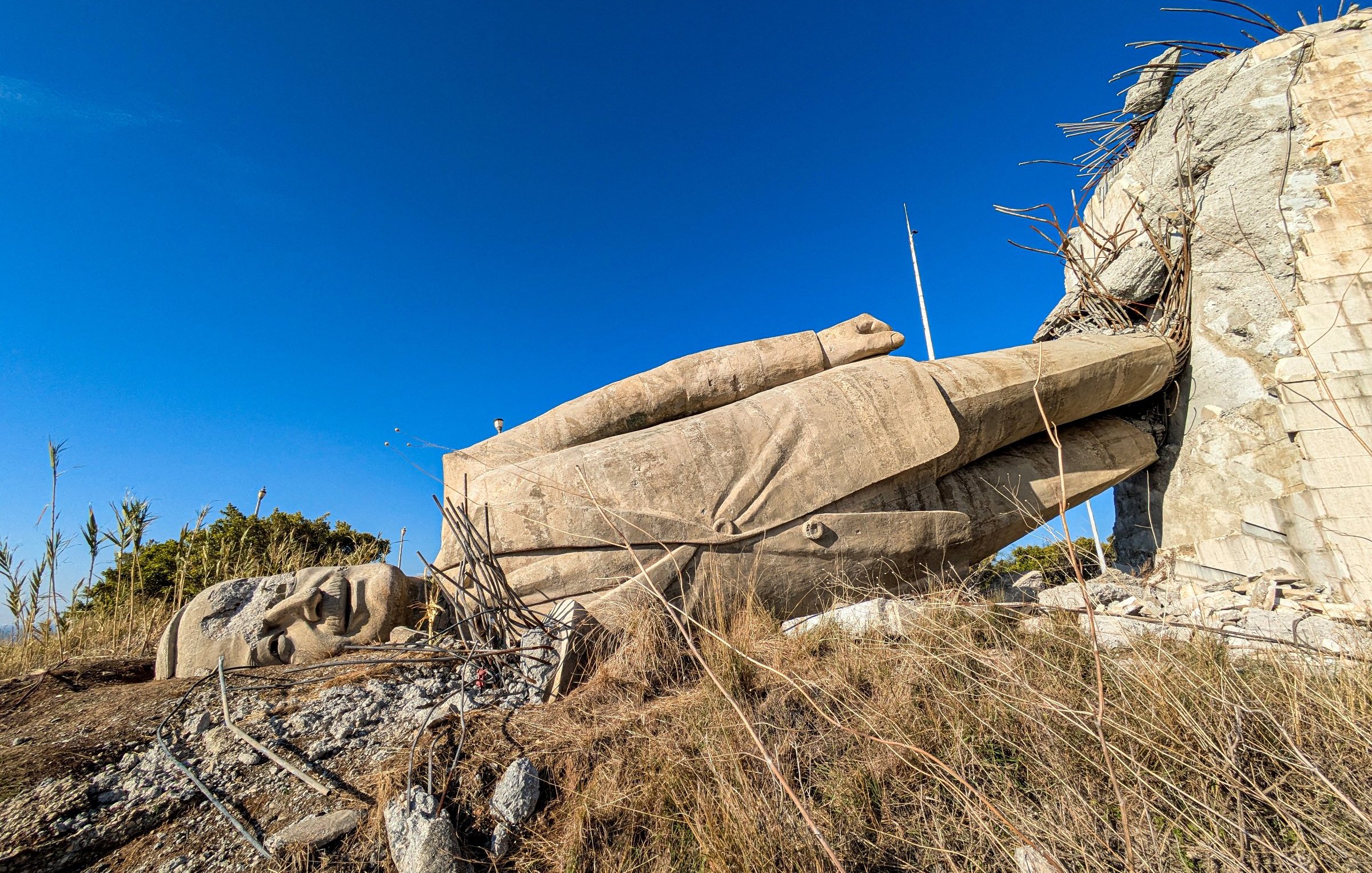 The image shows a large, partially fallen stone statue lying on its side against a rocky background, surrounded by sparse vegetation and a clear blue sky.