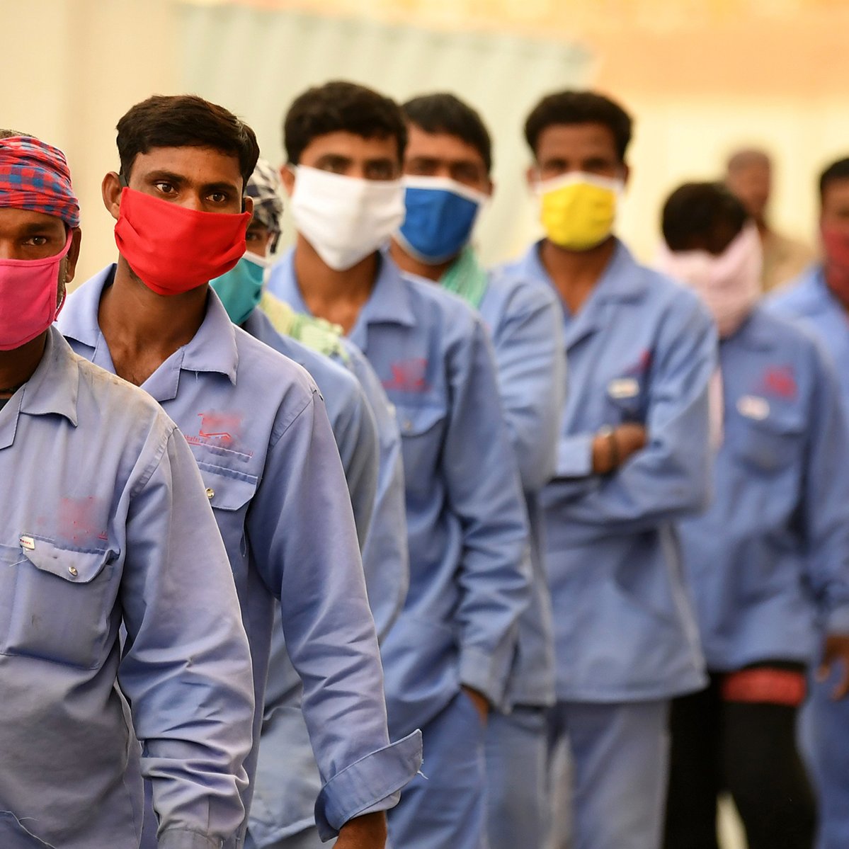 En la imagen se puede observar a un grupo de hombres alineados, todos ellos usando mascarillas de diferentes colores. Visten uniformes azules y parecen estar en un entorno al aire libre. La fila de personas avanza hacia algún destino, y el ambiente sugiere que podría ser una situación laboral o de espera. Algunos de los hombres llevan pañoletas en la cabeza, lo que indica posibles medidas de protección o identificación.