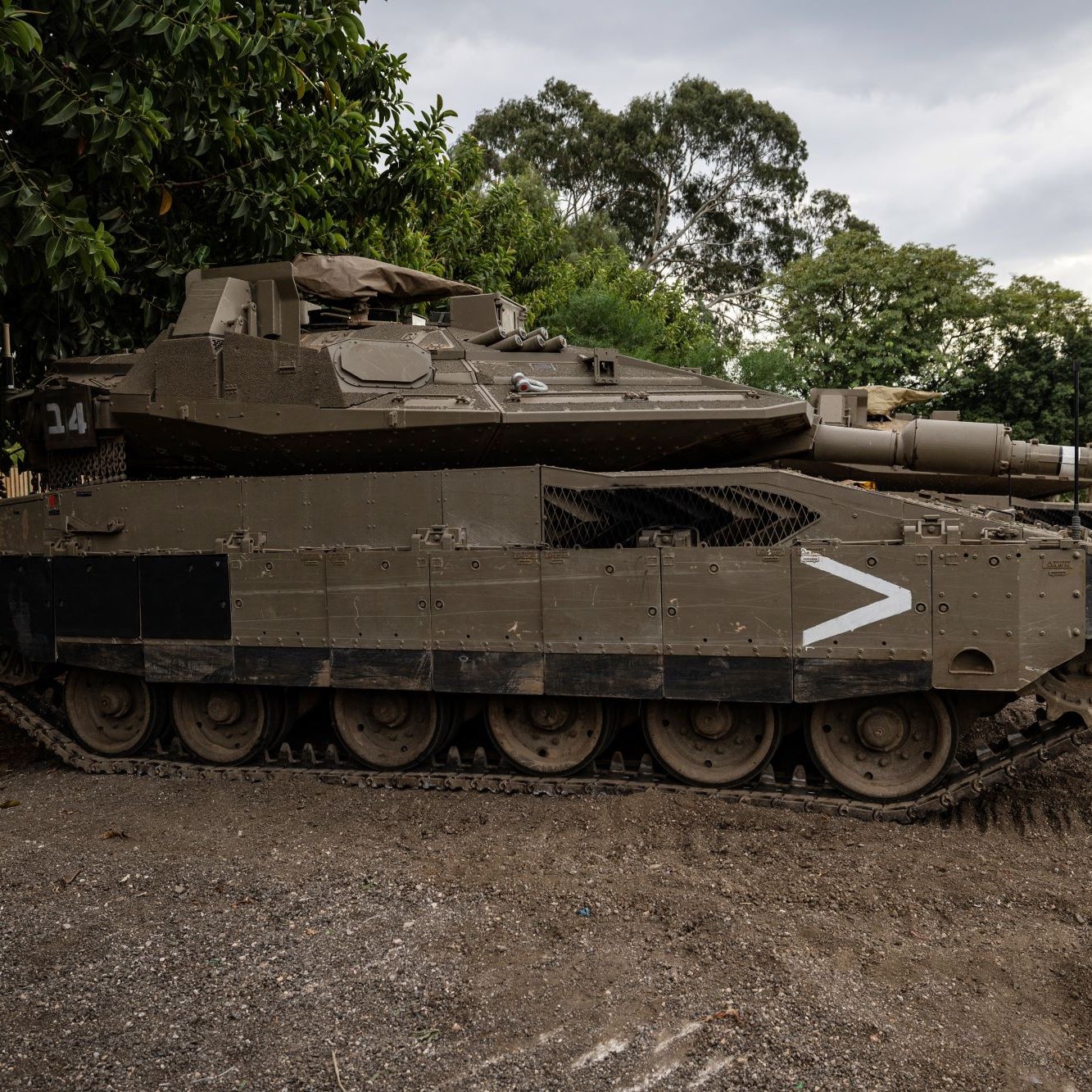 L'image montre un char d'assaut stationné sur un terrain. Le véhicule est de couleur verte foncée et possède des marques de camouflages. Il est équipé d'une tourelle avec un canon pointé vers l'avant. En arrière-plan, on peut voir des arbres et un ciel nuageux, ce qui donne une ambiance militaire à la scène. Le char semble robuste et bien conservé, prêt pour une utilisation éventuelle.