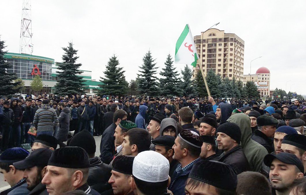 L'image montre une grande foule rassemblée dans une rue, probablement lors d'une manifestation ou d'un rassemblement public. Les participants semblent en majorité masculins et portent différents types de couvre-chefs. Certains tiennent des drapeaux, et l'atmosphère semble mobilisée. En arrière-plan, on peut voir des bâtiments et des arbres, ce qui donne un aperçu de l'environnement urbain. Le ciel est nuageux, ce qui pourrait indiquer un temps frais.