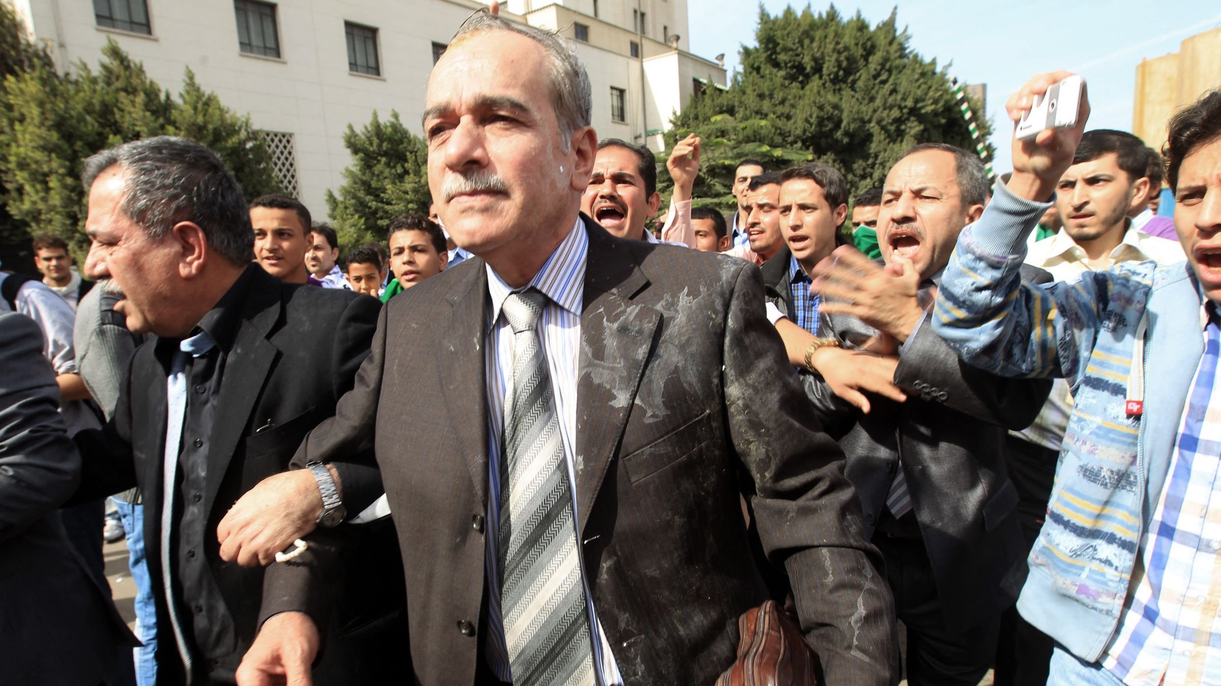 L'image montre un groupe de personnes dans une scène dynamique et animée. Au centre, un homme en costume, portant une cravate, semble marcher avec détermination, bien qu'il ait des éclaboussures sur son vêtement. Il est entouré de plusieurs autres individus qui semblent agités et expriment des émotions fortes, peut-être en criant ou en gesticulant. L'arrière-plan montre un environnement urbain avec des bâtiments et des arbres. L'atmosphère semble chargée, suggérant une situation de tension ou de protestation.