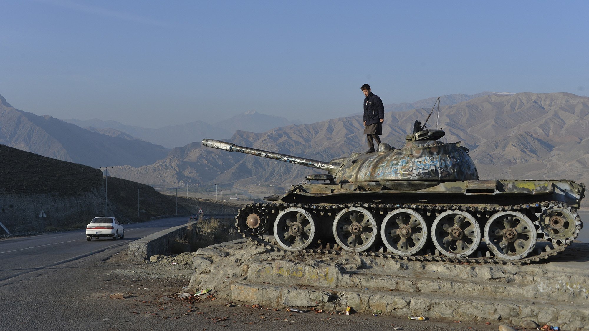 در این تصویر، یک تانک قدیمی با رنگ و زنگ‌زدگی مشاهده می‌شود که در یک نقطه مرتفع قرار دارد و فردی بر روی آن ایستاده است. پس‌زمینه شامل کوه‌های بلند و آسمان صاف است. در سمت راست تصویر، یک اتومبیل در حال حرکت در جاده‌ای کوهستانی دیده می‌شود. این صحنه نمایانگر تلاقی تاریخ و مناظر طبیعی است.