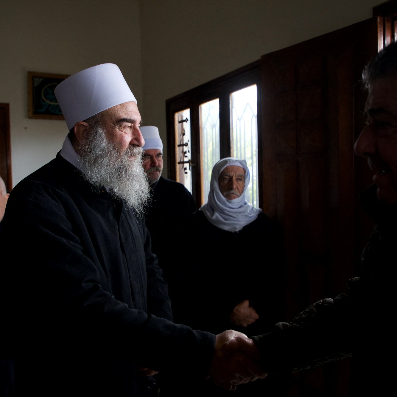 Group of men, some with white hats and beards, shaking hands and wearing dark clothing in a room with windows.
