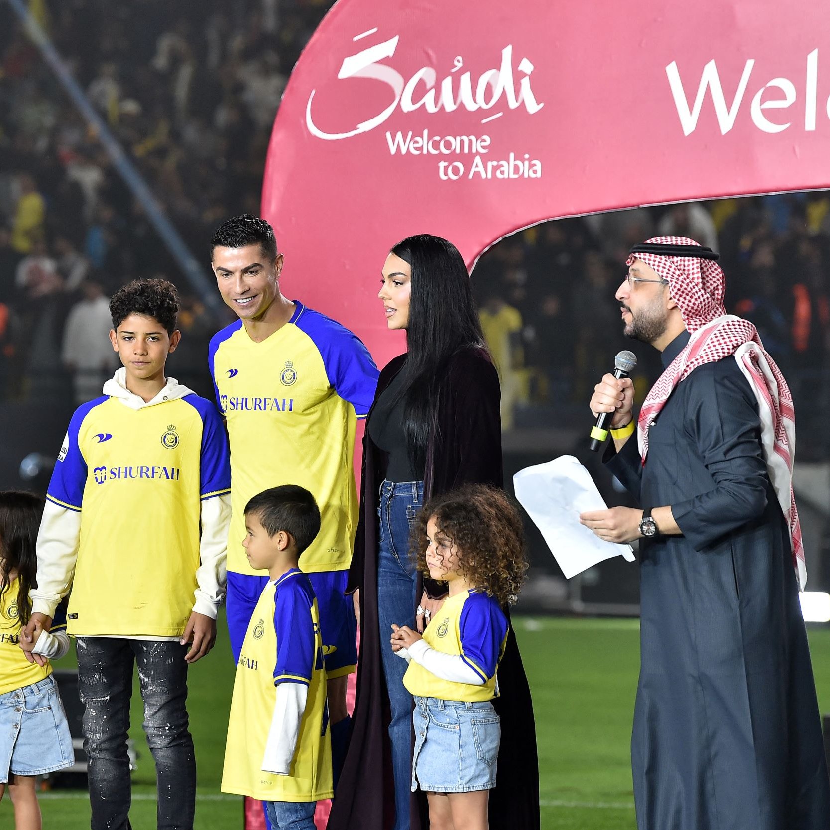 در این تصویر، یک رویداد ورزشی در حال برگزاری است. در وسط تصویر، کریستیانو رونالدو، بازیکن معروف فوتبال، همراه با چند کودک و یک زن ایستاده است. آن‌ها لباس‌های تیمی با رنگ زرد و آبی پوشیده‌اند. در پس‌زمینه، جمعیتی از طرفداران به وضوح دیده می‌شوند. همچنین، یک نفر در حال صحبت در میکروفون است و دو نفری که سمت چپ تصویر قرار دارند، به نظر می‌رسد که مجری برنامه هستند. به نظر می‌رسد که این رویداد به معرفی یا خوشامدگویی به رونالدو مربوط می‌شود. یک بنر بزرگ با نوشته "Welcome to Arabia" نیز در تصویر دیده می‌شود.