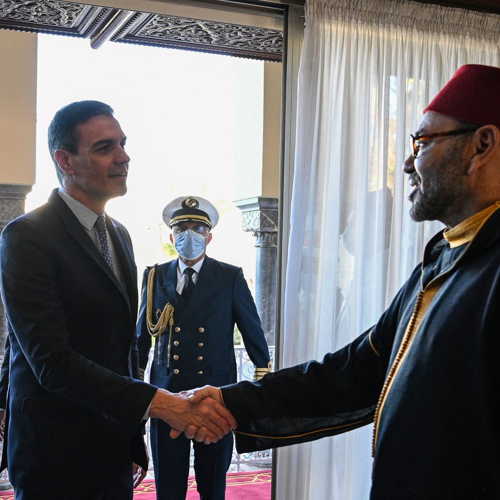 The image depicts a meeting between two men. One man, dressed in a suit, is shaking hands with the other man, who is wearing traditional attire, including a red fez. In the background, a military figure stands in uniform, observing the handshake. The setting appears to be an elegantly decorated room with large windows and elaborate architectural details.