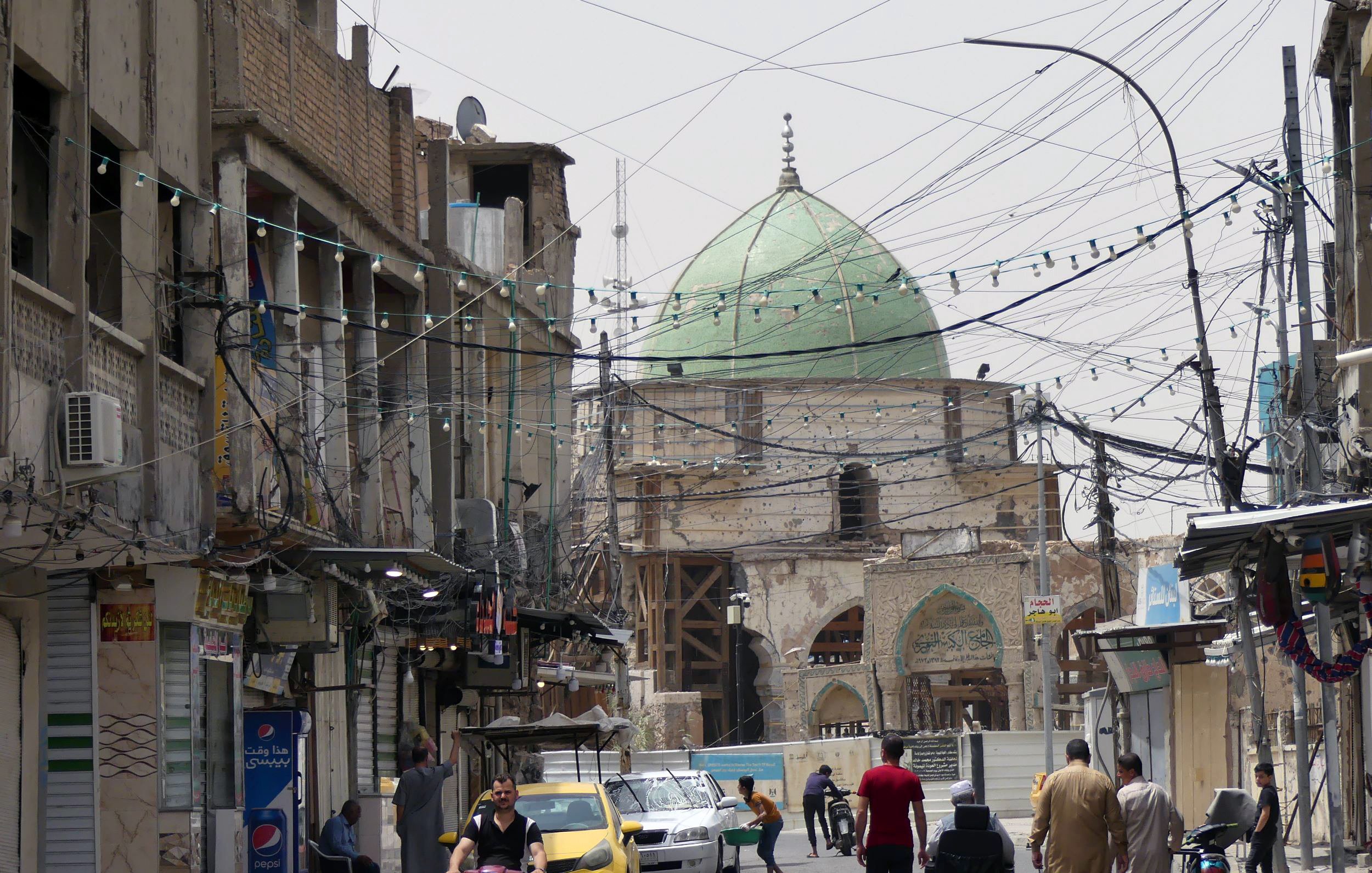 الصورة تظهر شارعًا في منطقة حضرية، حيث يتواجد عدد من الأشخاص يسيرون في اتجاهات مختلفة. في الخلفية، يظهر مسجد ذو قبة خضراء، مما يعكس الطابع المعماري المحلي. الشارع مليء بوسائل النقل، حيث توجد دراجة نارية وسيارة صفراء. بالإضافة إلى ذلك، تبرز الأسلاك الكهربائية المعلقة في الهواء والمحال التجارية على جوانب الطريق، مما يضفي على المشهد طابعًا حيويًا ونابضًا بالحياة.