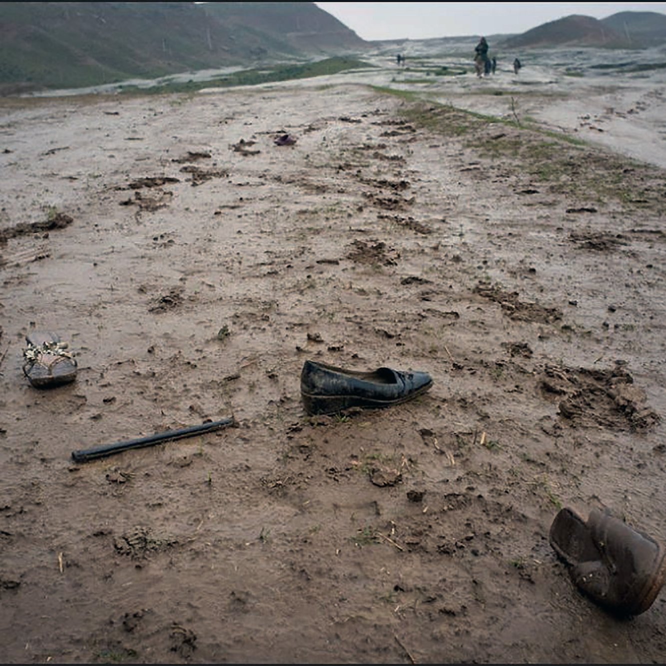 La imagen muestra un paisaje desolado y húmedo, con un terreno cubierto de barro y hierba. Hay varios zapatos dispersos por el suelo, uno de ellos es un zapato de tacón negro, otro parece ser un zapato de niño y hay un objeto más que podría ser un bastón o un palo. El ambiente parece gris y melancólico, y la presencia de barro sugiere que ha llovido recientemente. En el fondo, se pueden distinguir algunas figuras humanas que caminan a lo lejos.