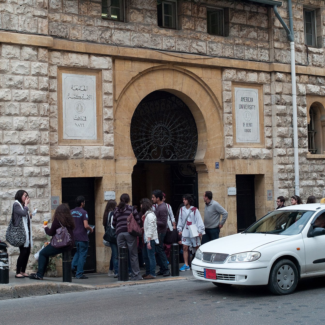الصورة تظهر مدخل جامعة معمارية مميزة، حيث يمكن رؤية واجهة المبنى التي تتكون من حجر باهت وأعمدة مميزة. أمام المدخل، يوجد مجموعة من الطلاب أو الأشخاص في صف، مما يدل على أنهم قد يكونون في انتظار التسجيل أو الدخول. إلى جانب المدخل، توجد دراجة نارية ومركبة تسير على الطريق، مما يعطي انطباعاً عن الحياة اليومية والحركة في المنطقة.背景وضح صورة جوًا من النشاط والاهتمام بالتعليم.