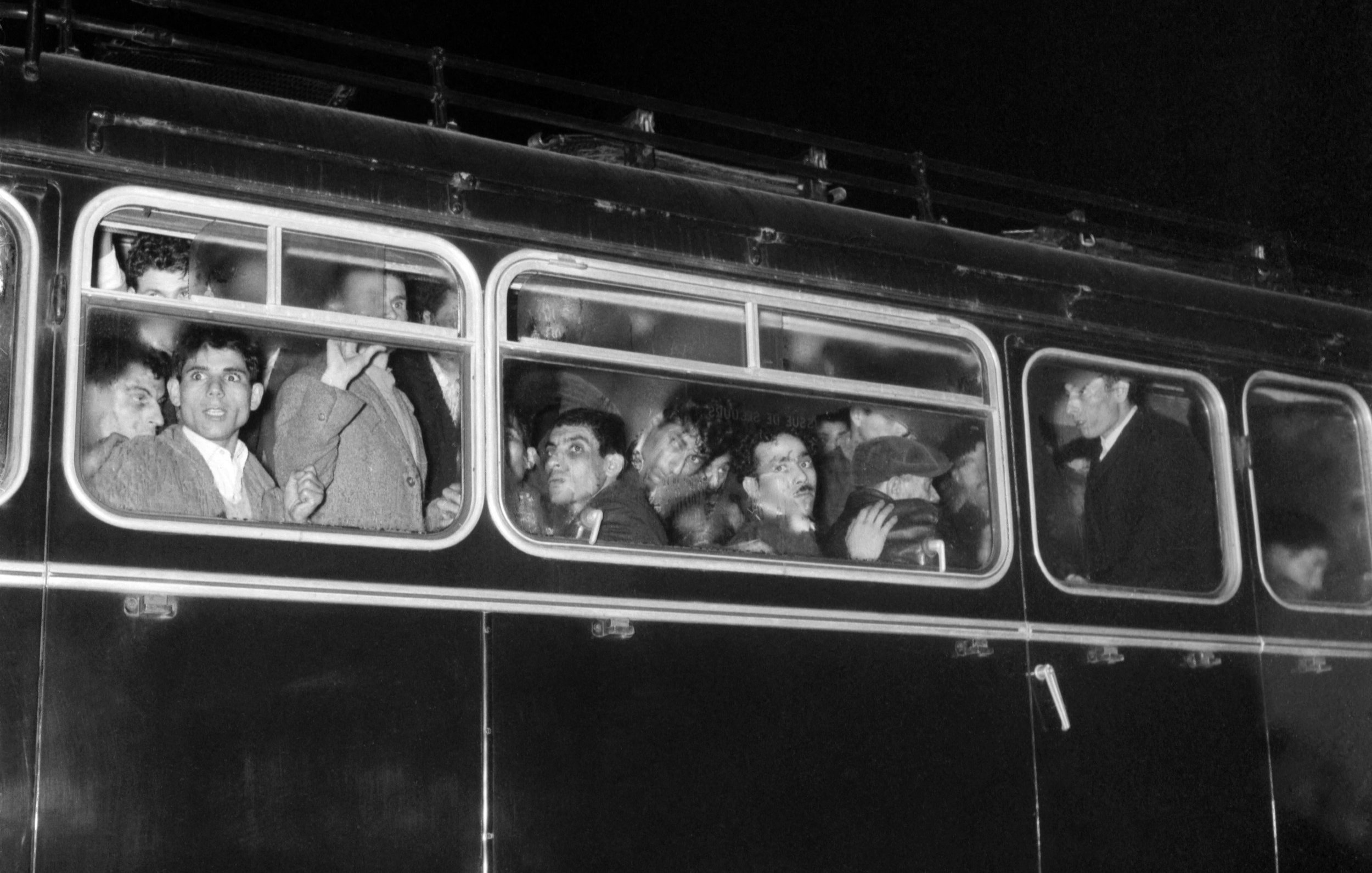 تصویر نشان‌دهنده یک اتوبوس است که در شب در حال حرکت است. در داخل اتوبوس، چندین نفر وجود دارند که از پنجره‌ها به بیرون نگاه می‌کنند. چهره‌های آن‌ها به وضوح قابل مشاهده است و به نظر می‌رسد در حال گفتگو یا ابراز احساساتند. طراحی اتوبوس و فضای اطراف، حالتی تاریخی و نوستالژیک دارد. رنگ تصویر به سیاه و سفید است که بر جذابیت آن افزوده و حس زمان گذشته را القا می‌کند.