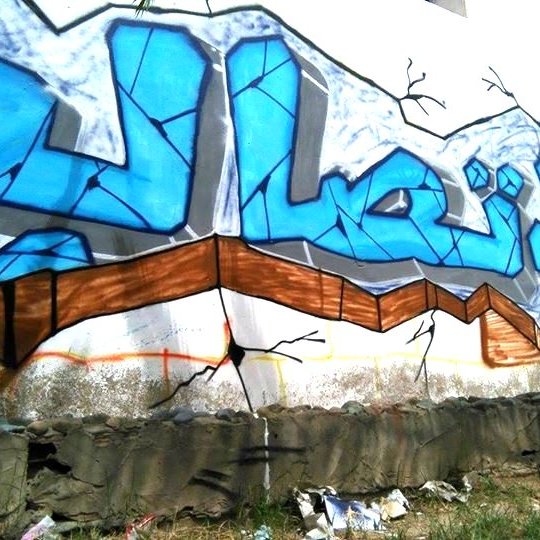 L'image montre un mur de graffiti avec des lettres stylisées en bleu. Le fond est légèrement cassé, ce qui donne l'impression que le mur est en train de se fissurer. Le mot écrit semble être en arabe, avec des motifs et des contours en noir qui accentuent le design. En bas, on peut apercevoir des déchets et de l'herbe, ce qui montre que l'art se trouve dans un environnement urbain.