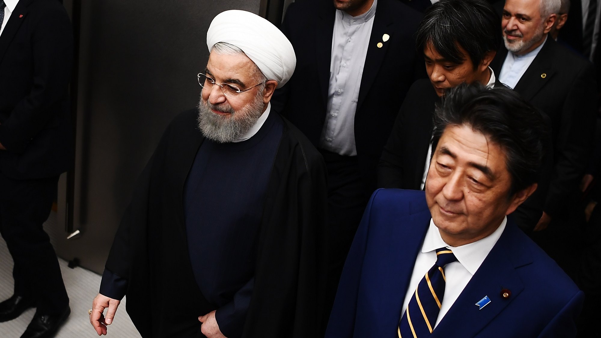 The image shows two prominent political figures walking side by side. The person on the left is wearing a traditional clerical outfit, including a white turban and long black robe, while the person on the right is dressed in a formal suit with a tie. They appear to be engaged in conversation or moving through a venue, accompanied by several other individuals in the background. The setting appears to be formal and possibly involves diplomatic discussions or a meeting.