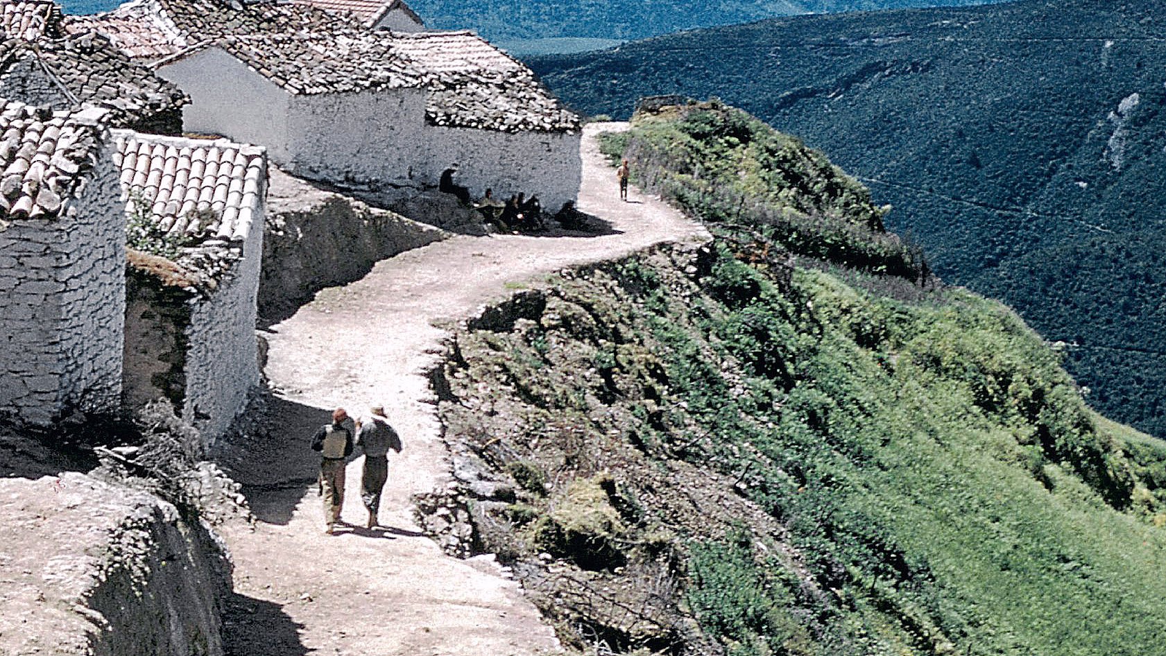 L'image montre un paysage rural avec un sentier serpentant le long d'une colline. De chaque côté du chemin, on peut voir des maisons blanches avec des toits en tuiles. Deux personnes marchent sur le sentier, profitant de la vue. En arrière-plan, des montagnes verdoyantes s'élèvent, créant une atmosphère paisible et naturelle. Le ciel est clair et contribue à l'ambiance sereine de la scène.