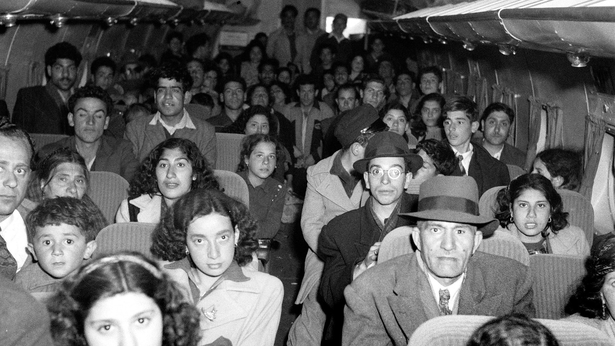 الصورة تُظهر مجموعة كبيرة من الأشخاص داخل طائرة، حيث يبدو أن معظمهم يجلسون في مقاعدهم. هناك تنوع في الأعمار، بدءًا من الأطفال إلى البالغين. يمكن رؤية تعبيرات مختلفة على وجوه الركاب، حيث يبدون جميعًا في حالة انتظار أو استعداد للسفر. تصميم الطائرة بسيط، مع نوافذ صغيرة فوق مقاعد الركاب. الأزياء تبدو تقليدية، مما يعطي انطباعًا عن زمن قديم.