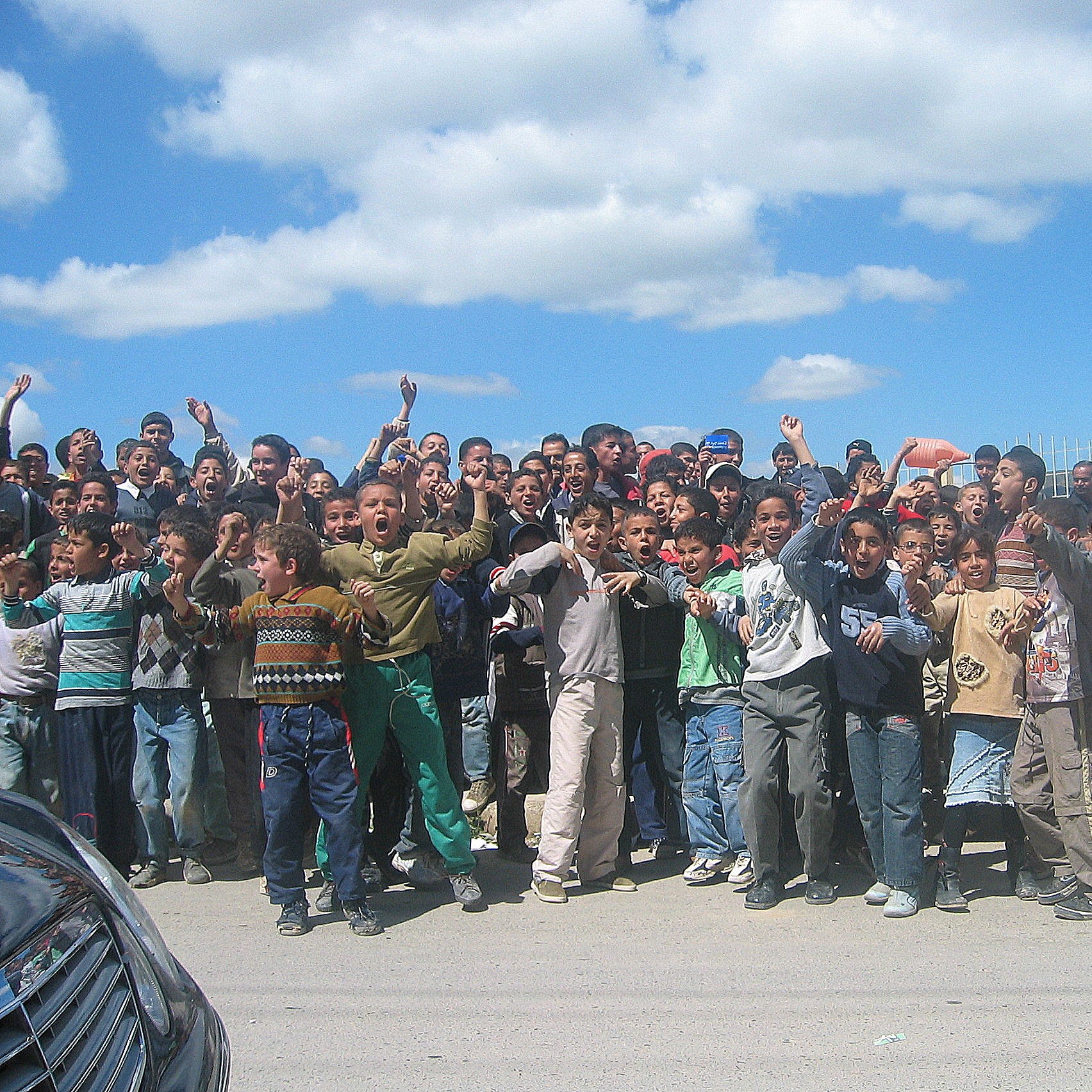 تظهر في الصورة مجموعة كبيرة من الأطفال والشباب يتجمعون في مكان واحد، يبدو أنهم متحمسون ويقومون بالتفاعل مع شيء ما، ربما يجتمعون لمتابعة حدث أو نشاط معين. السماء في الخلفية زرقاء مع بعض السحب، والمكان يبدو كأنه في منطقة حضرية. يرتدي الأطفال ملابس متنوعة، وتعكس تعبيراتهم الحماس والإثارة.