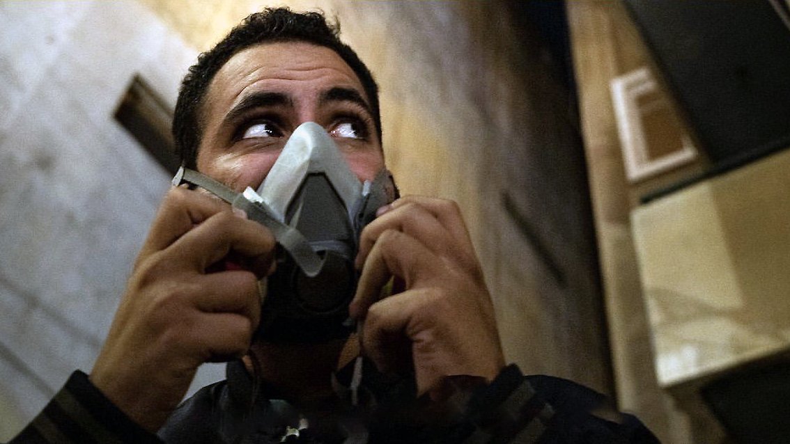 L'image montre un homme qui porte un masque à gaz. Son visage exprime une certaine vigilance, et il semble regarder autour de lui. L'environnement est urbain, avec des murs et des bâtiments en arrière-plan. L'éclairage donne une ambiance sombre et intense à la scène.