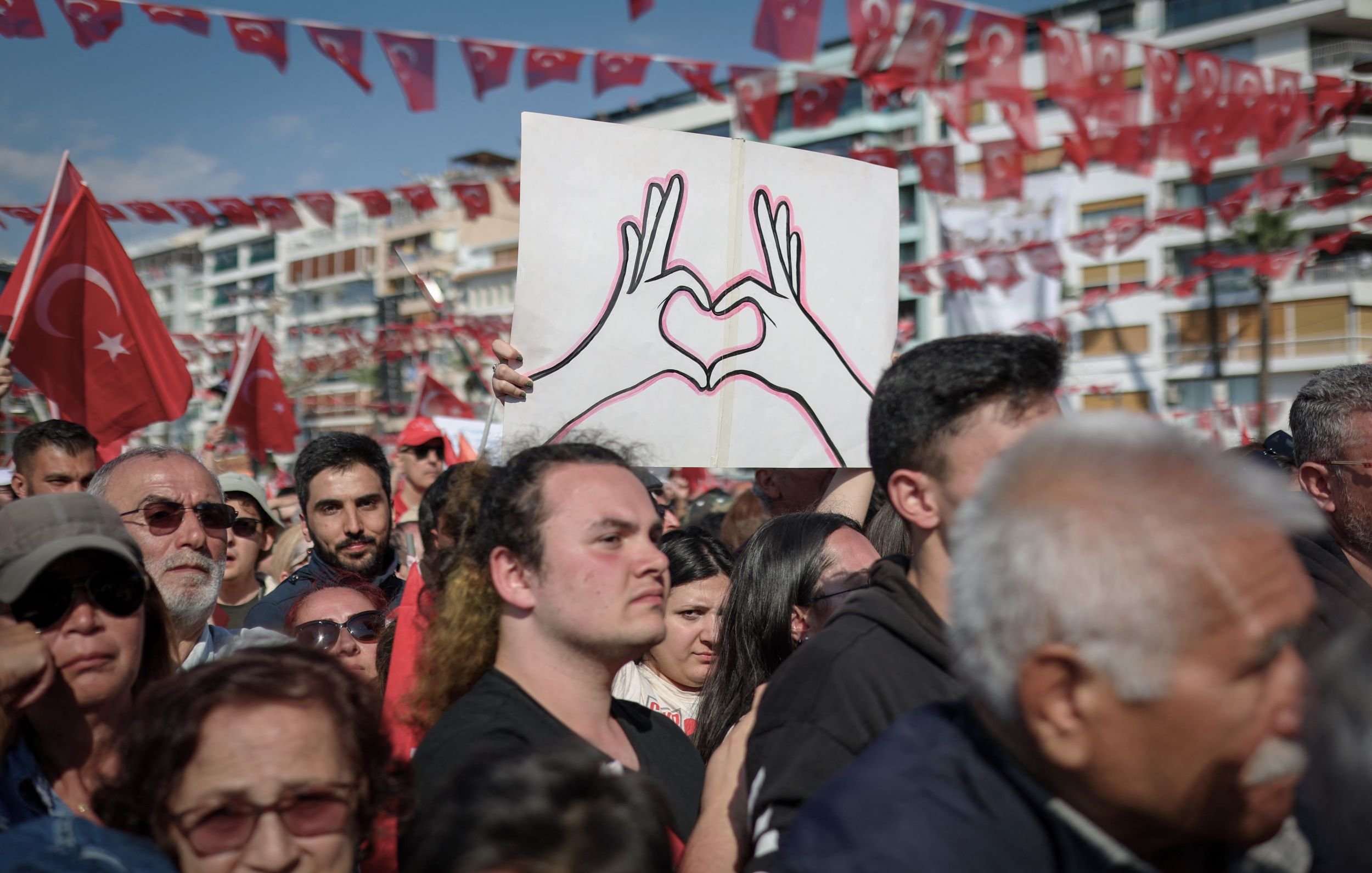 L'immagine mostra una grande folla riunita in un evento all'aperto, probabilmente una manifestazione o una celebrazione. Alcuni partecipanti sostengono bandiere turche, che sono di un rosso acceso, mentre un uomo in primo piano è visibile con una maglietta nera e guarda verso la telecamera. Dietro di lui, c'è un cartello che rappresenta due mani che formano un cuore, simbolo di amore o unità. L'atmosfera sembra essere festosa e piena di energia, con molte persone che condividono un momento di comunità.