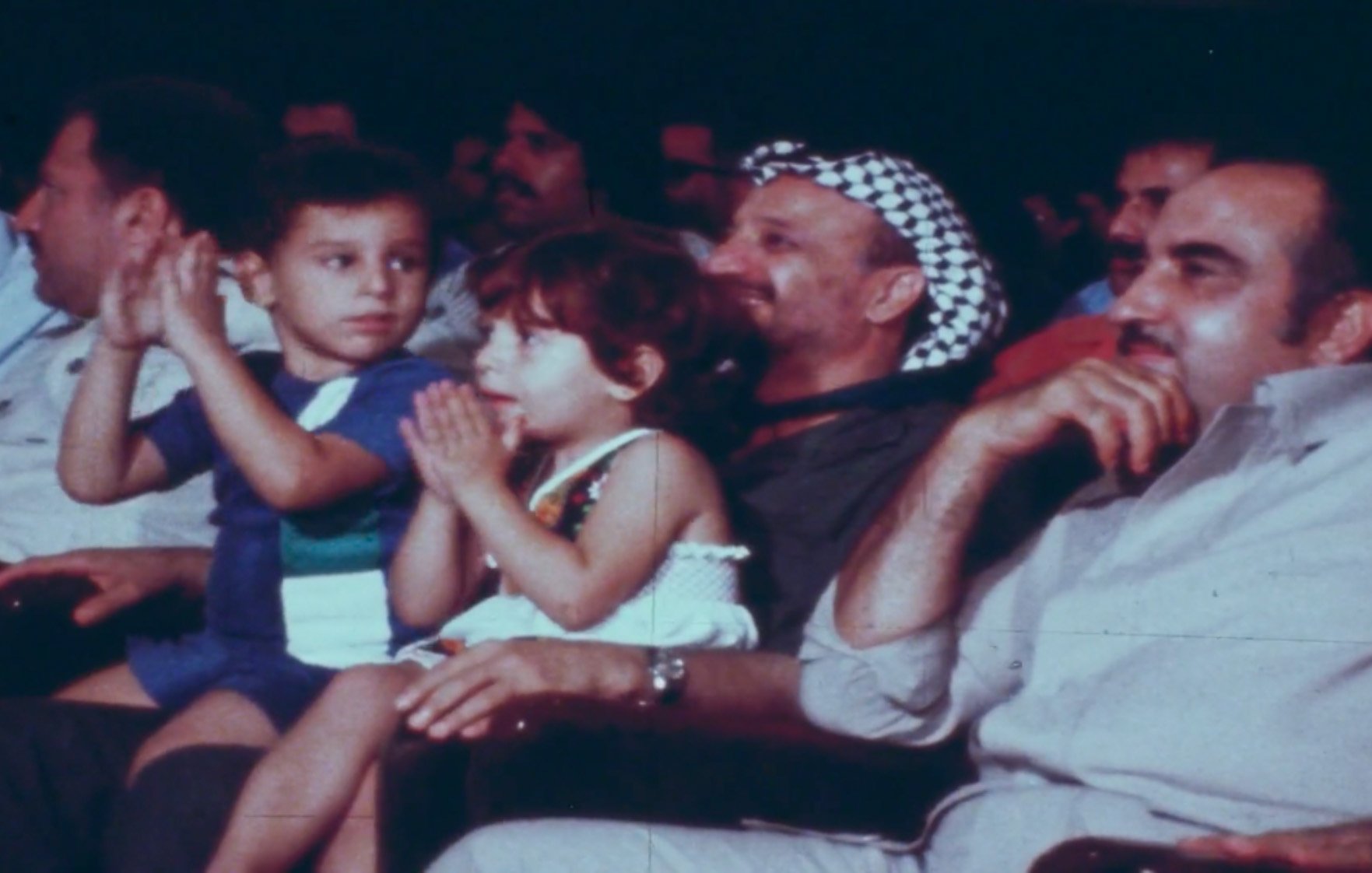 في الصورة مجموعة من الأشخاص يجلسون في قاعة، ومن بينهم طفلان يجلسان أمام رجل يرتدي كوفية. يبدو أن الجميع يستمع أو يشاهد شيئًا ما في الفعالية. الأطفال يظهرون تفاعلهم بالتصفيق أو الاستمتاع بالعرض. الرجال في الصورة يبدون مهتمين ومسترخين. الجو يبدو احتفاليًا أو حيويًا.