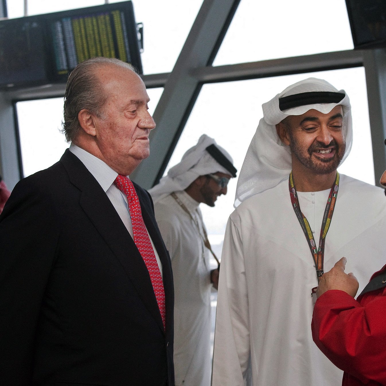 The image shows a gathering of three men engaged in conversation. Two of them are dressed in traditional attire, typical of Middle Eastern cultures, with one wearing a white kandura and the other in a black and white headdress. The third man is in formal Western attire with a suit and a red tie. The background suggests an airport or a similar setting, indicated by the large windows and digital displays. The expressions of the men appear to be friendly and engaged in a dialogue.
