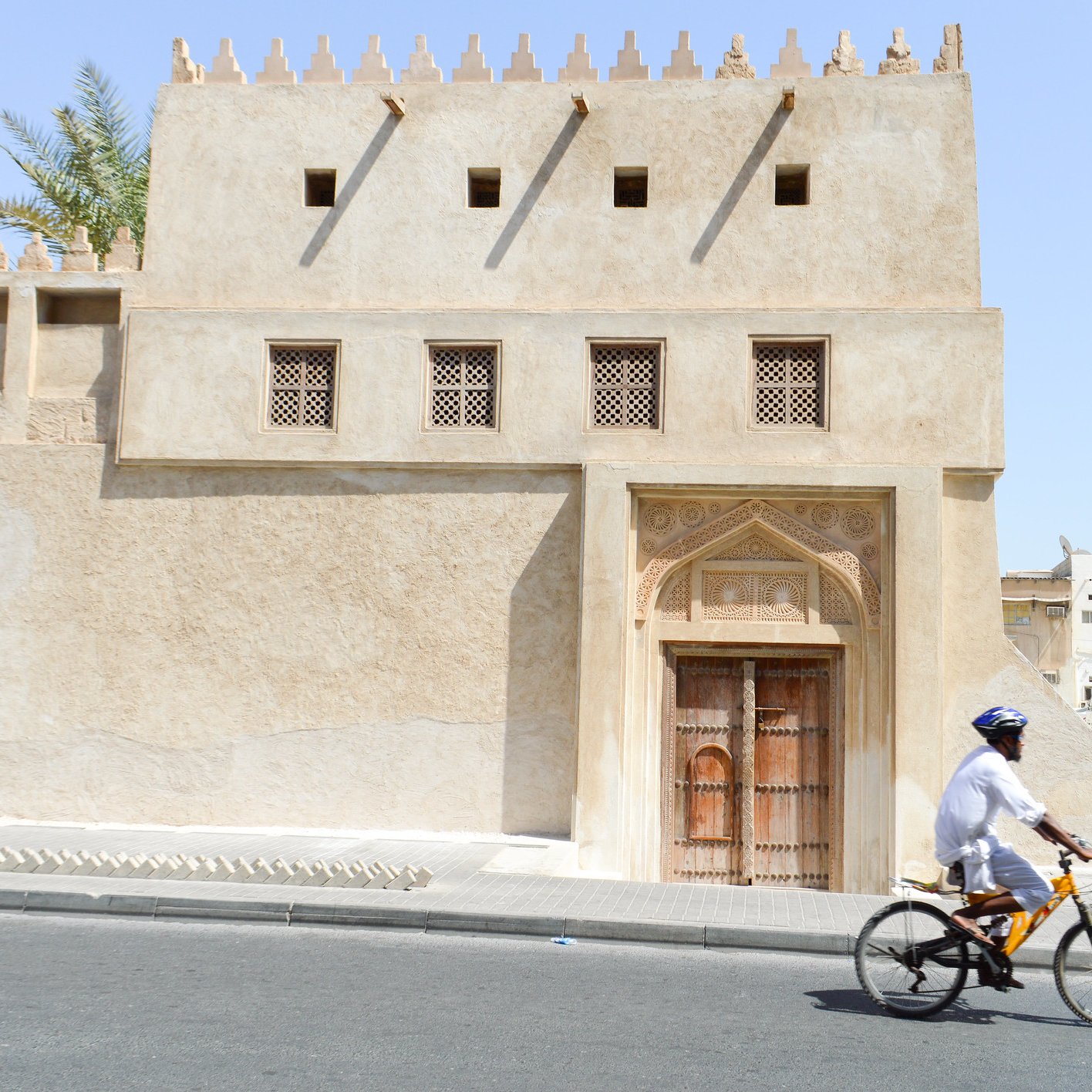 La imagen muestra un edificio de arquitectura tradicional, con paredes de color beige y detalles ornamentales en la fachada. La puerta principal es de madera tallada, y hay ventanas con rejillas decorativas. Al lado de la construcción, un ciclista vestido con ropa ligera y un casco pedalea por una calle pavimentada. El ambiente parece cálido, lo que sugiere un clima árido, típico de ciertas regiones. En el fondo, se pueden observar otros edificios y estructuras, que contribuyen a la sensación de un entorno urbano.