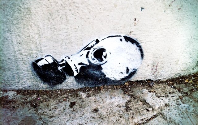 L'image montre un graffiti représentant une tête de mort portant un masque à gaz. Ce dessin est peint en noir et blanc et est appliqué sur un mur, près du sol. Le contraste entre le noir et le blanc met en évidence les contours du masque et de la tête, créant une image saisissante et inquiétante. Le style est à la fois moderne et urbain, typique des œuvres de street art.