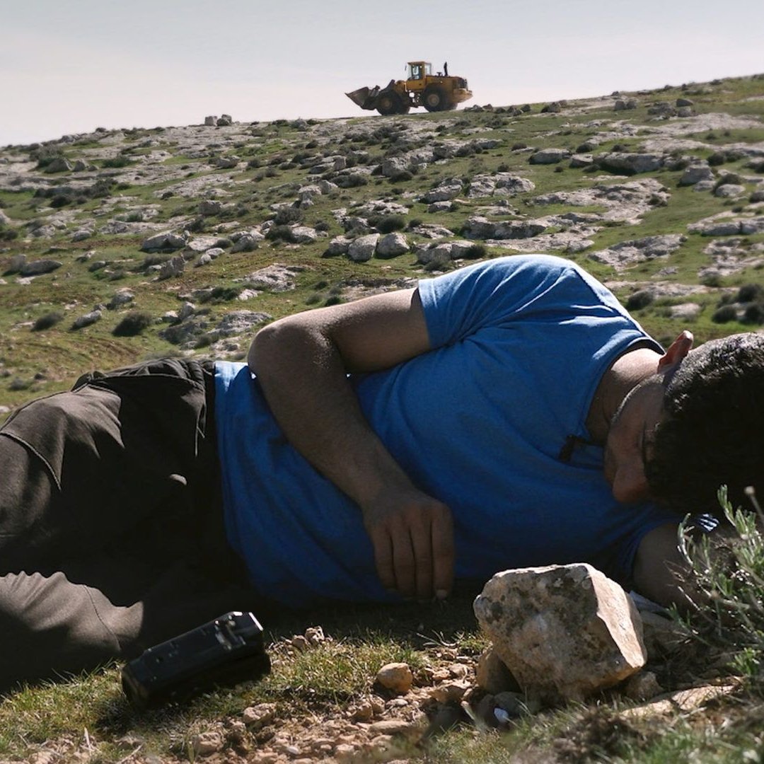 Nell'immagine si vede un uomo sdraiato su un prato verde, con il viso rivolto verso il terreno. Indossa una maglietta blu e pantaloni scuri. Accanto a lui ci sono alcune rocce. Sullo sfondo, si scorge un bulldozer che lavora su un terreno collinare. La scena trasmette un senso di tranquillità, ma anche di inquietudine a causa della presenza del macchinario che sottolinea il contrasto tra l'uomo e l'ambiente circostante.