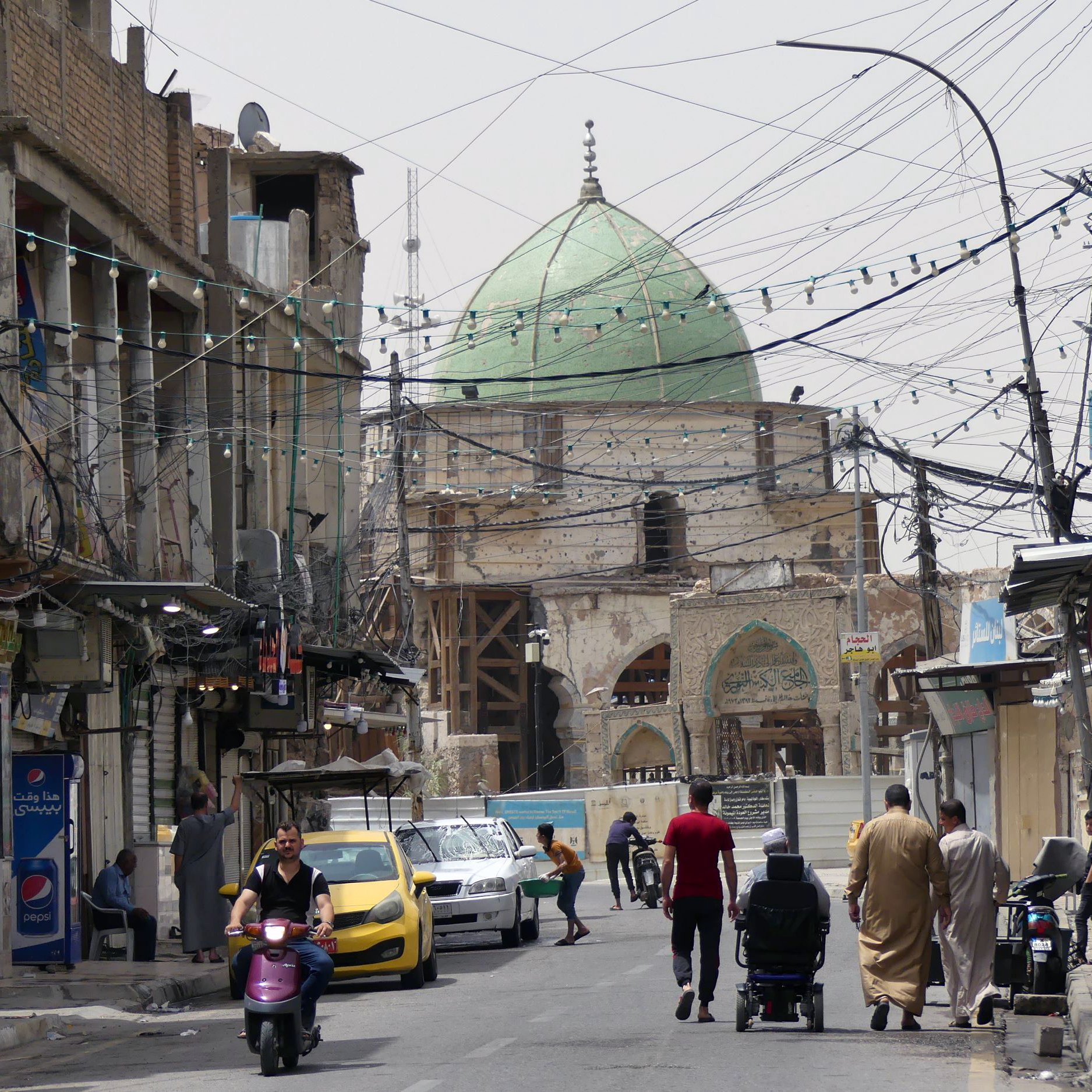 الصورة تظهر شارعًا في منطقة حضرية، حيث يتواجد عدد من الأشخاص يسيرون في اتجاهات مختلفة. في الخلفية، يظهر مسجد ذو قبة خضراء، مما يعكس الطابع المعماري المحلي. الشارع مليء بوسائل النقل، حيث توجد دراجة نارية وسيارة صفراء. بالإضافة إلى ذلك، تبرز الأسلاك الكهربائية المعلقة في الهواء والمحال التجارية على جوانب الطريق، مما يضفي على المشهد طابعًا حيويًا ونابضًا بالحياة.