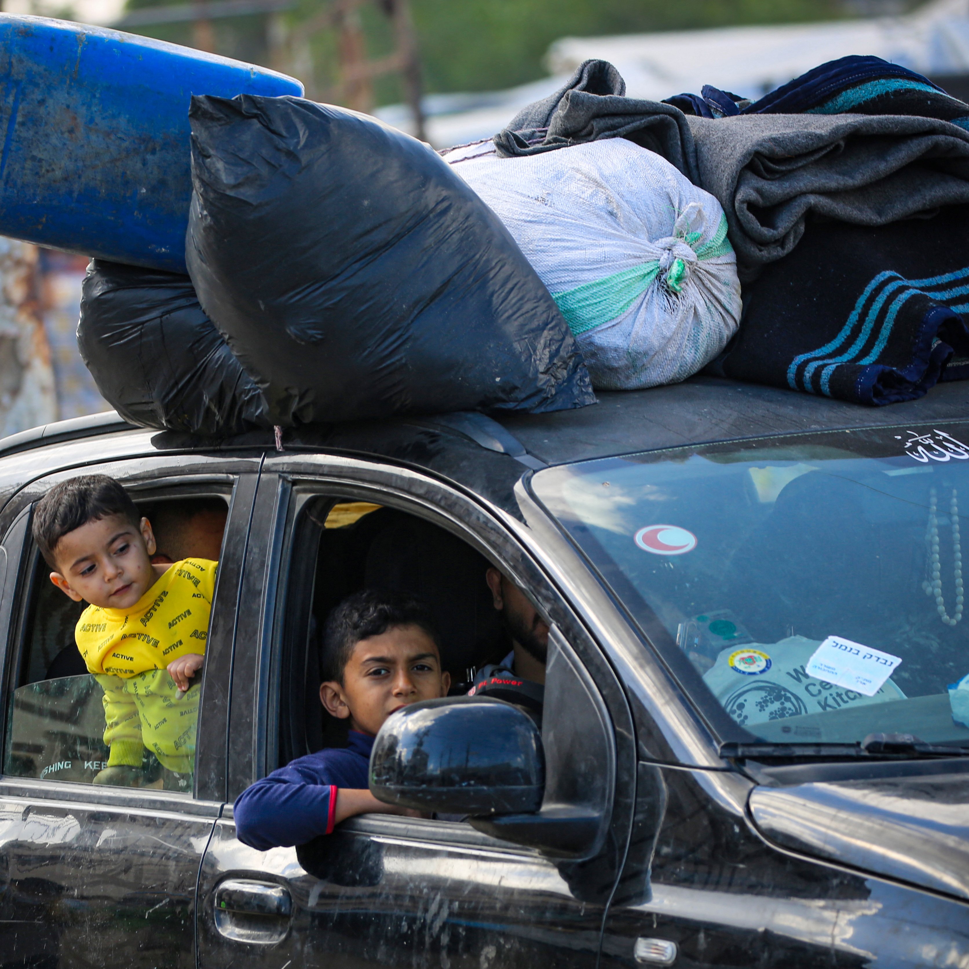 در این تصویر، یک خودرو دیده می‌شود که باری بر روی سقف آن قرار دارد. تعدادی کیسه و لباس به شکل نامنظم روی سقف خودرو گذاشته شده‌اند. درون خودرو، دو کودک نشسته‌اند؛ یکی از آن‌ها در حال نگاه کردن به بیرون از پنجره است و دیگری به سمت او توجه دارد. در پس‌زمینه، افرادی در حال حرکت هستند و فضایی شلوغ و شهری به نظر می‌رسد. وضعیت کلی تصویر نشان‌دهنده زندگی روزمره در یک محیط حاشیه‌ای است.